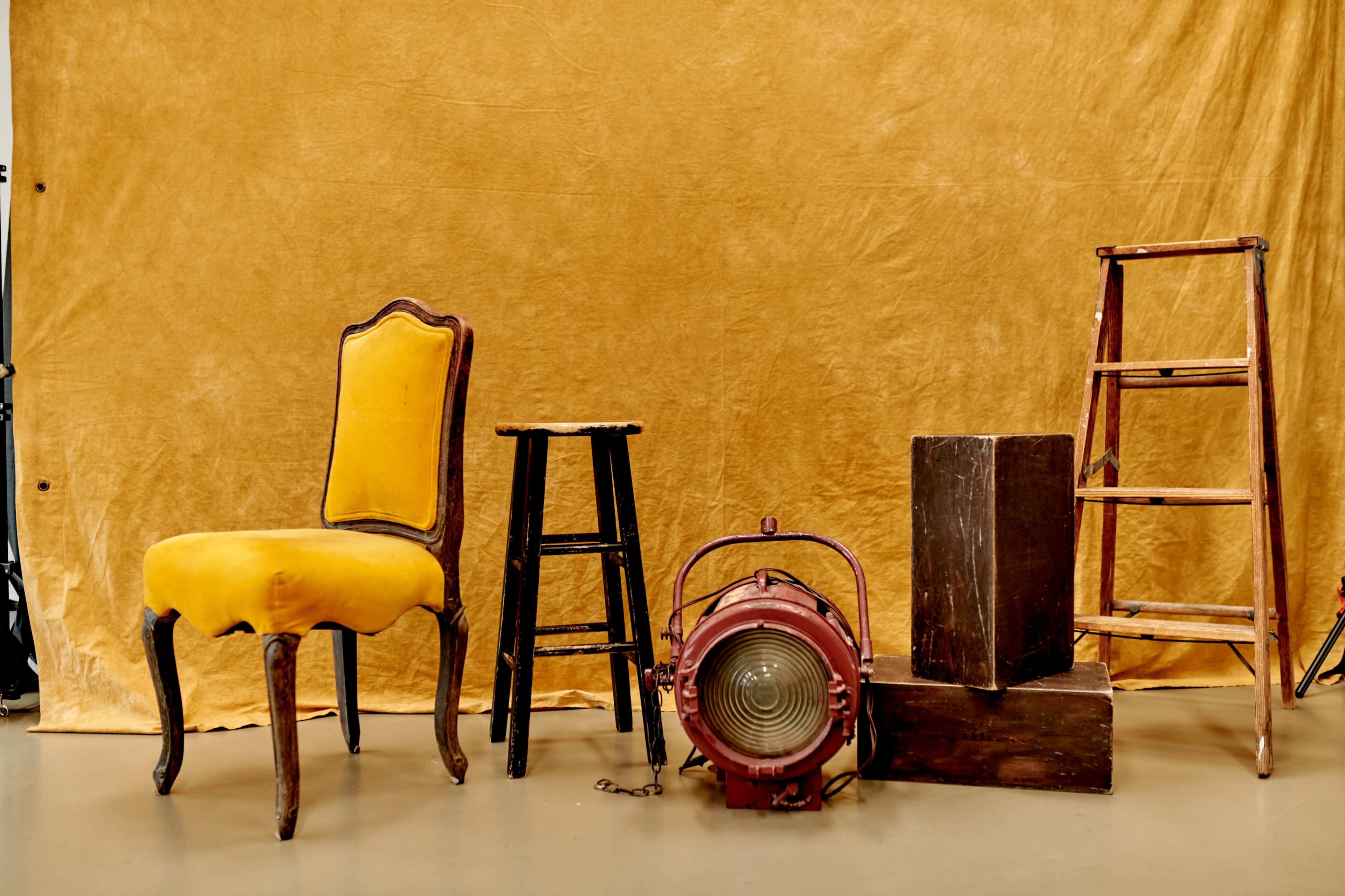 A yellow upholstered chair, a black stool, a vintage spotlight, a wooden box, and a step ladder arranged in front of a textured golden background.
