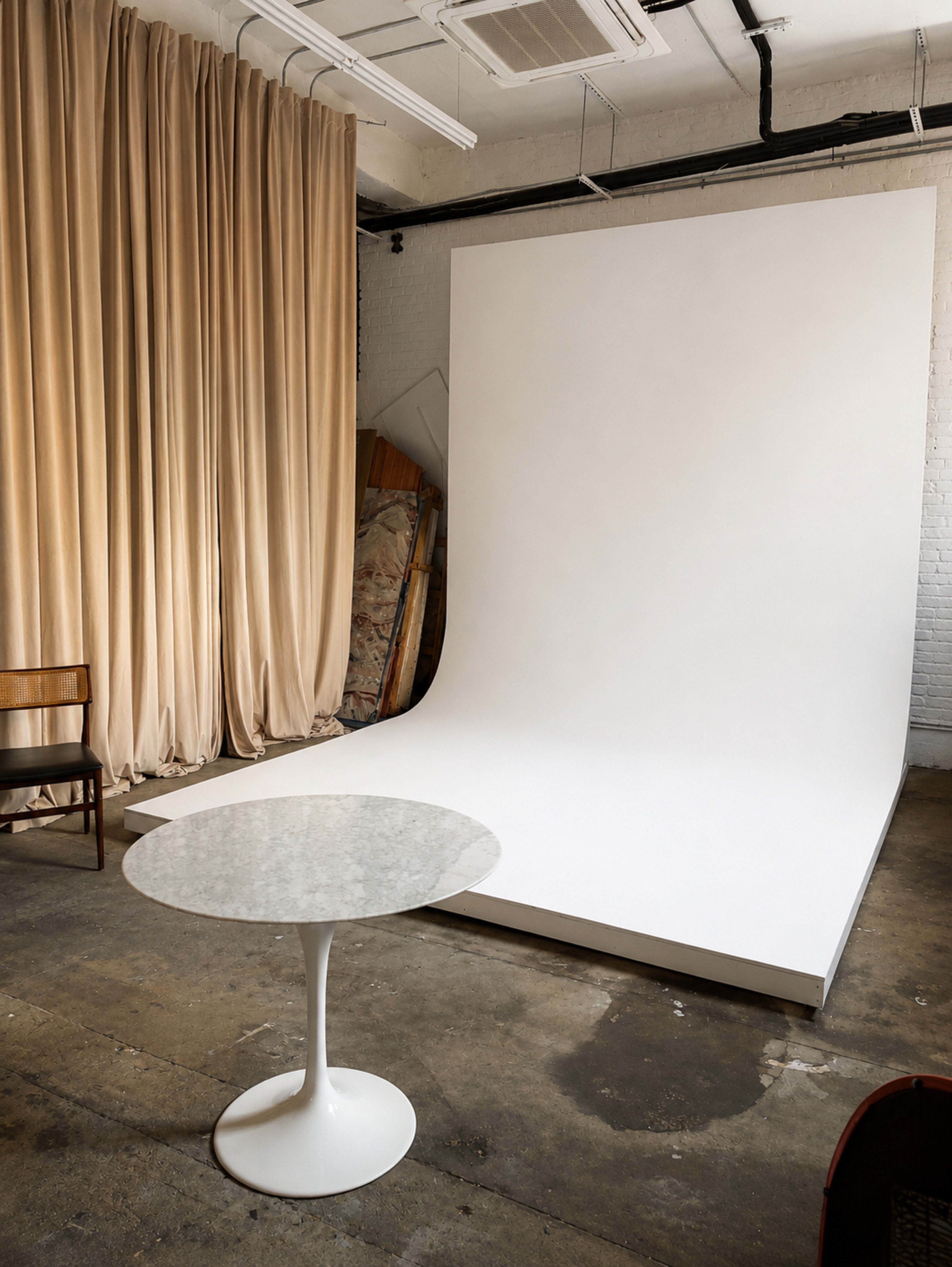 The image shows a minimalist photography studio featuring a large white backdrop, a circular white table, and a curtain.