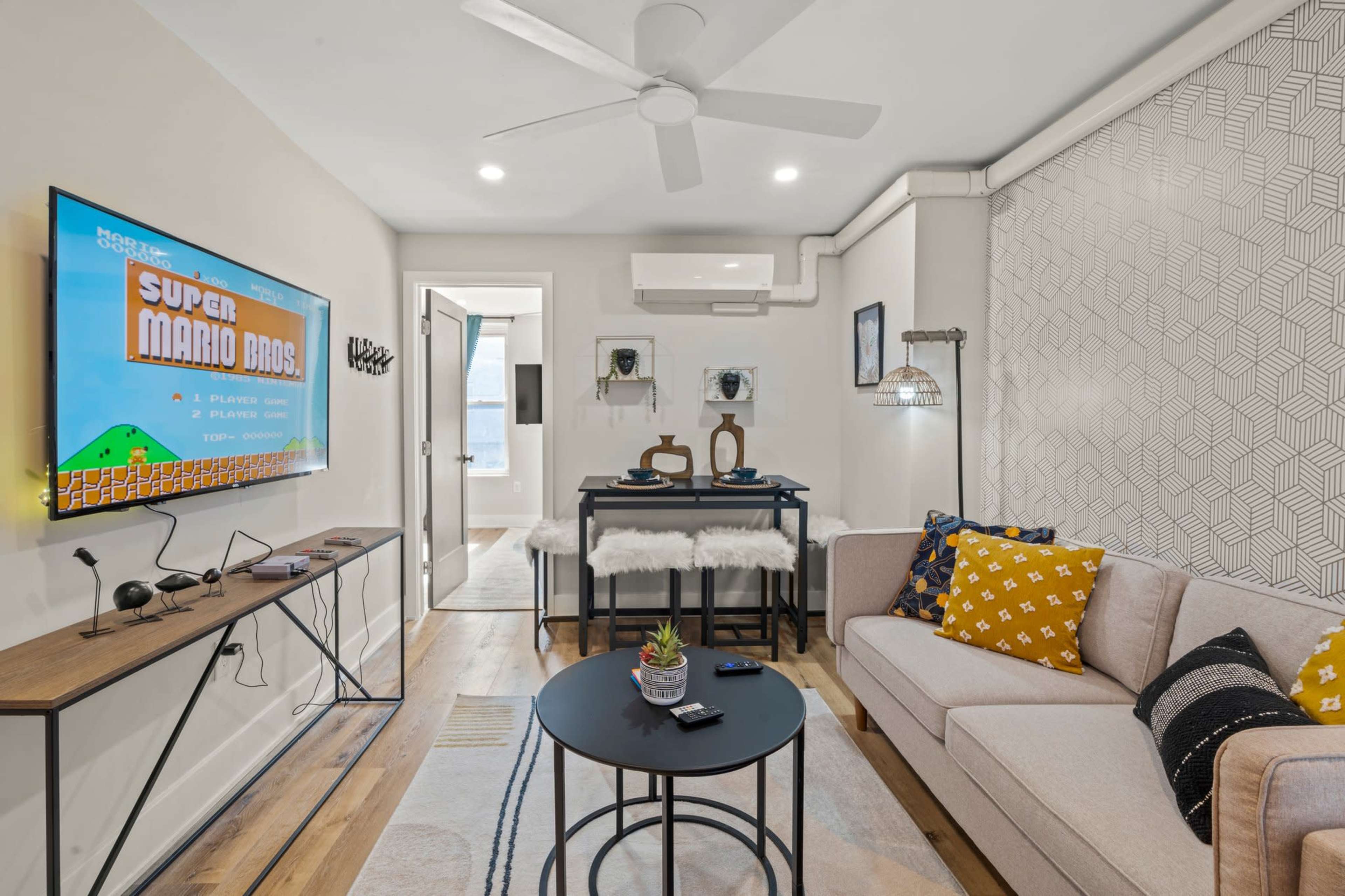 A modern living room featuring a wall-mounted TV displaying a classic video game, a coffee table, and a sofa adorned with decorative pillows.