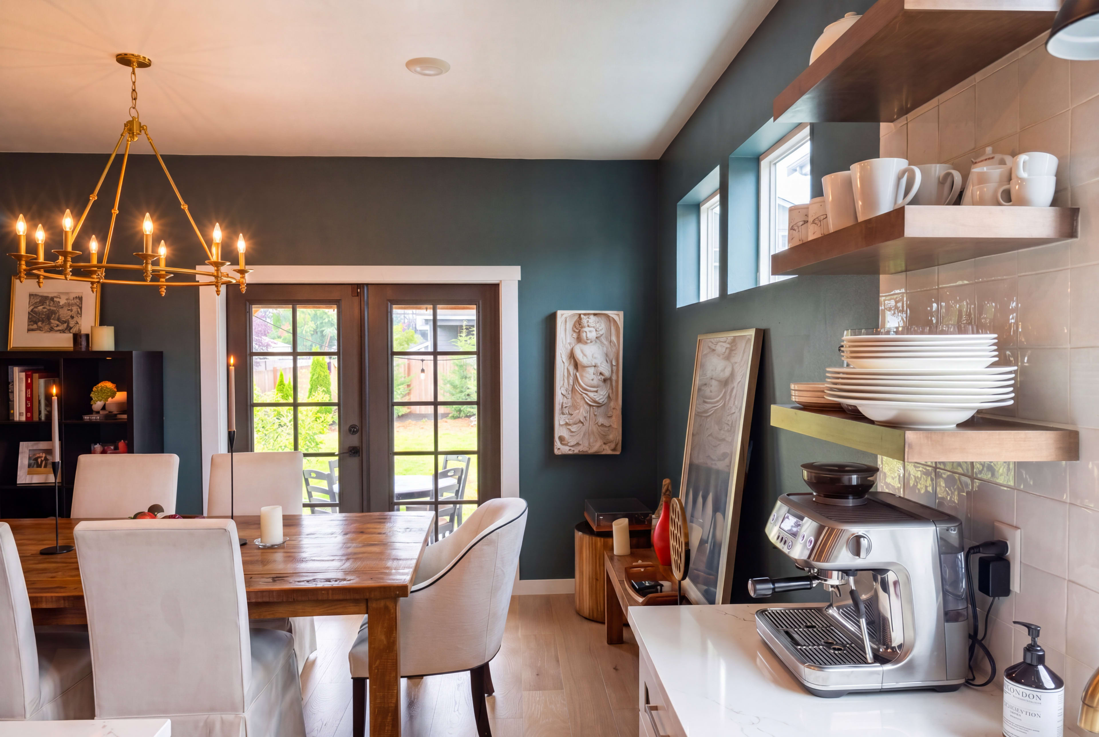 The image shows a modern dining area featuring a wooden table, white upholstered chairs, a chandelier, and open shelves displaying dishes next to a coffee machine, with large windows offering a view of a garden.