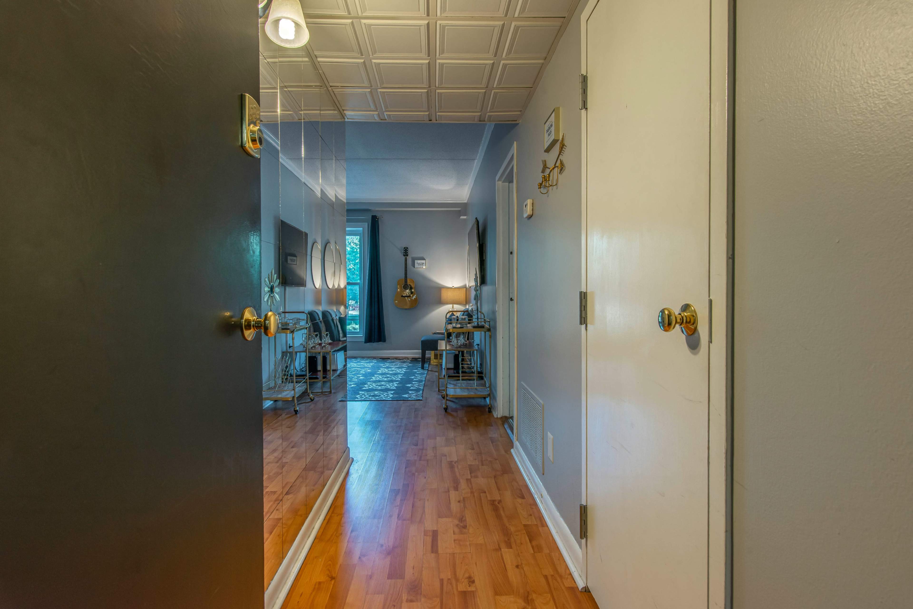 A hallway with a mirrored wall, leading to a living area with wooden flooring and a small table near the entrance.