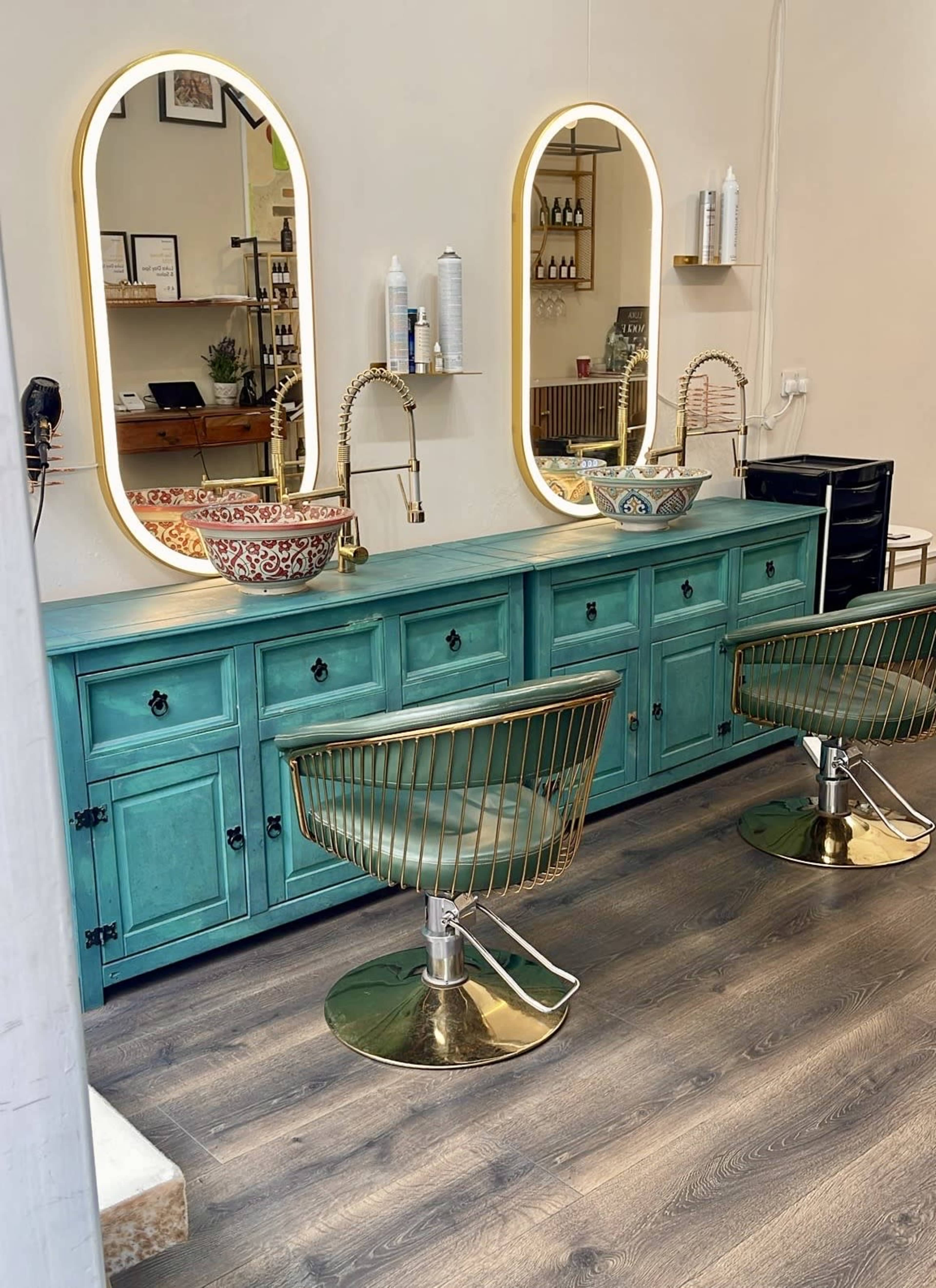 The image shows a modern salon interior featuring two stylish hairdressing chairs, a turquoise cabinet with washbasins, and large oval mirrors illuminated by warm lighting.