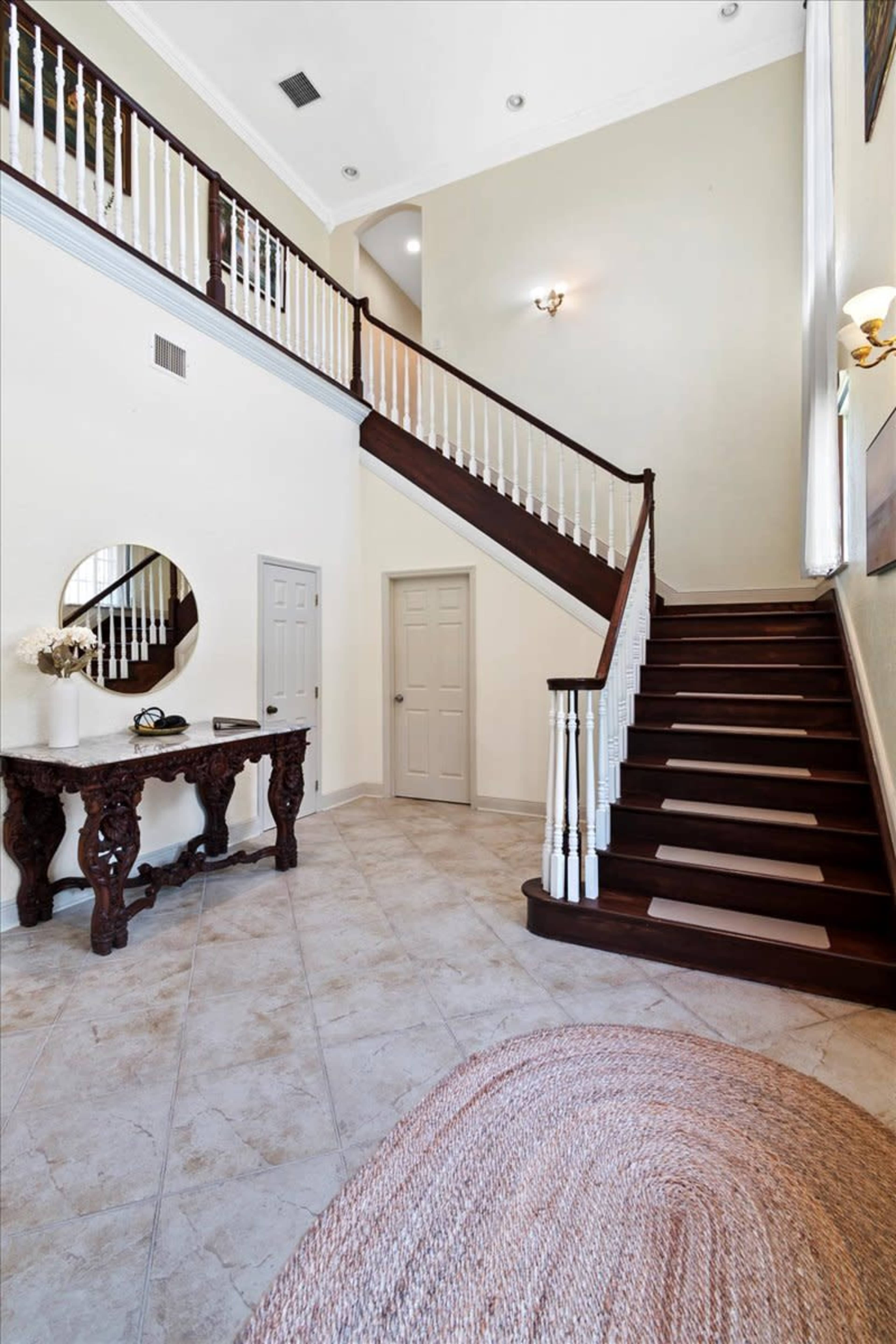 A spacious foyer with a staircase leading to an upper level, a round mirror on the wall, and a decorative table on the side.
