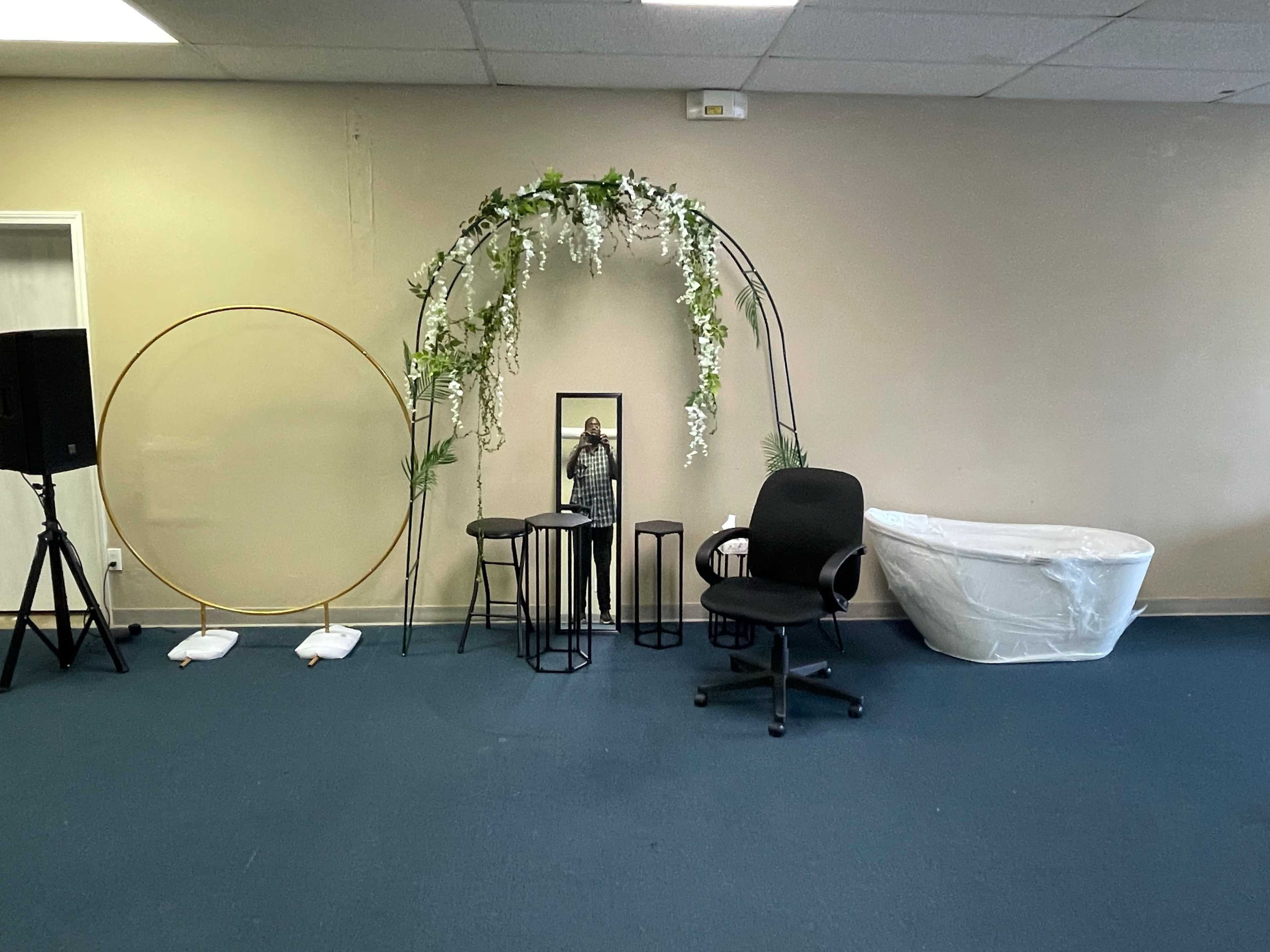 The scene features a minimalist indoor space with a decorative floral arch, a round hoop, a mirror, three black stools, an office chair, and a white bathtub against a plain wall.