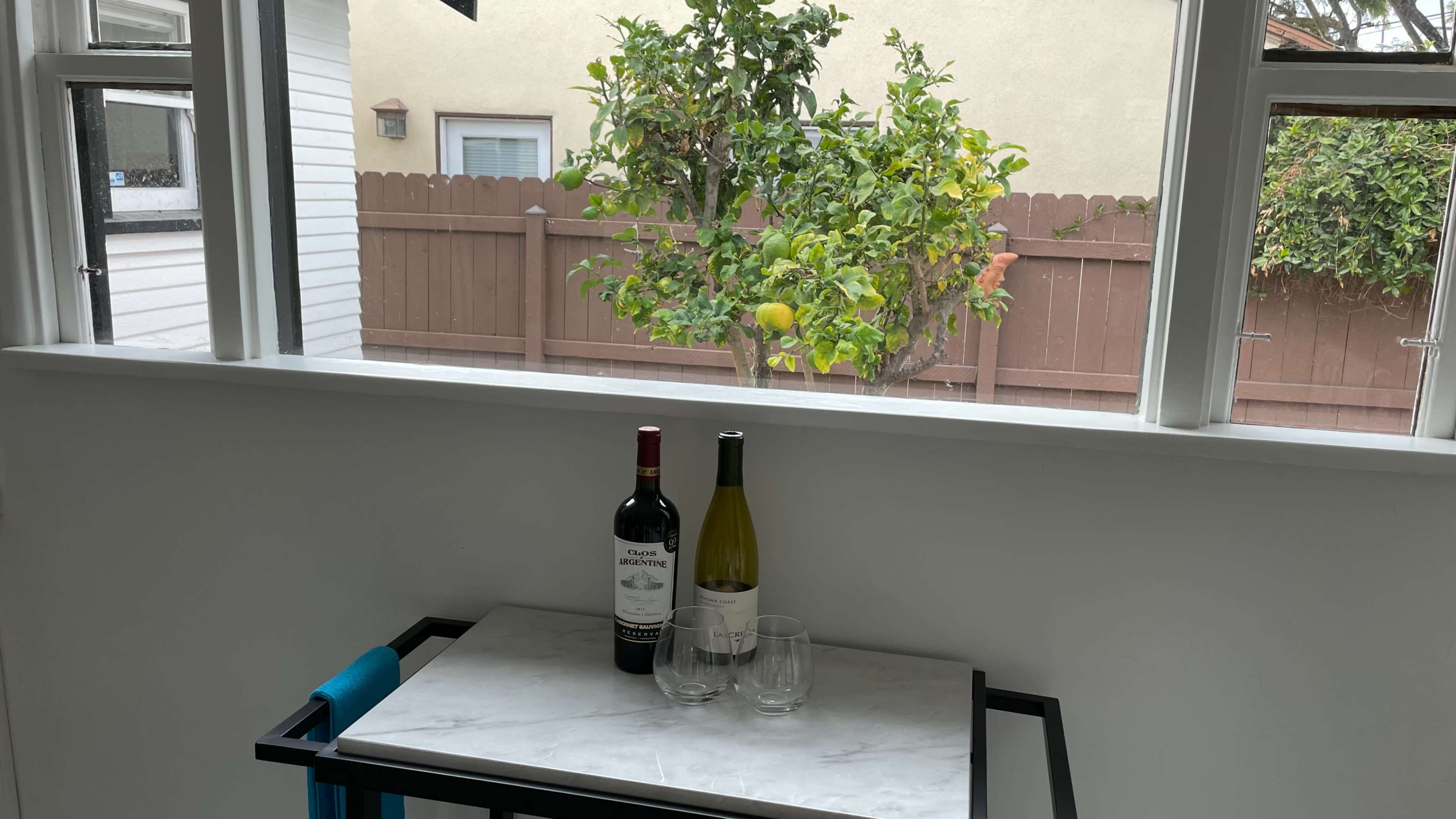 A small table with two bottles of wine and two glasses is positioned near a window overlooking a tree with green leaves and fruit in a fenced backyard.