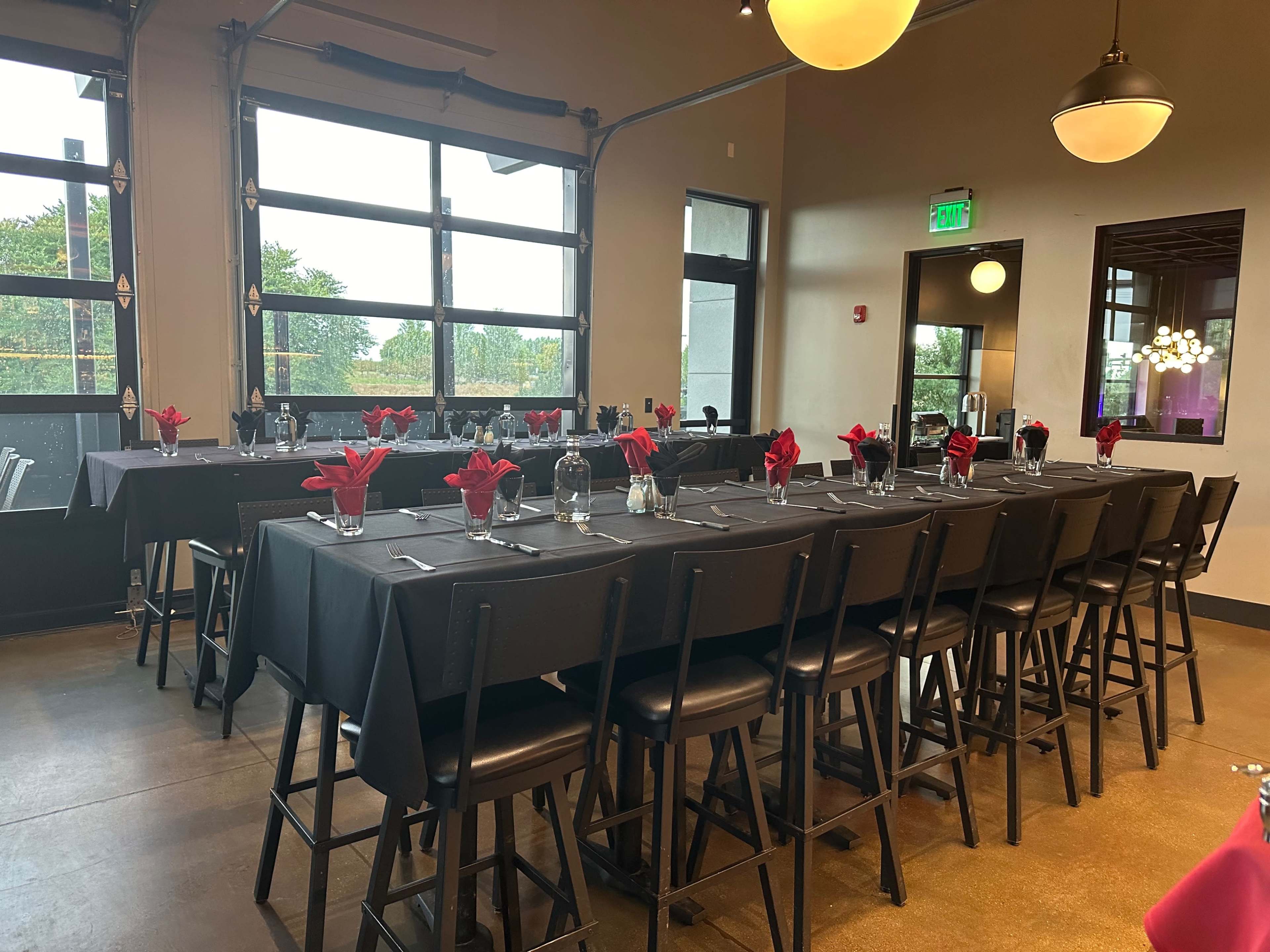 The image shows a dining setup featuring long tables covered with black tablecloths, each adorned with red napkins and glassware, in a venue with large windows.