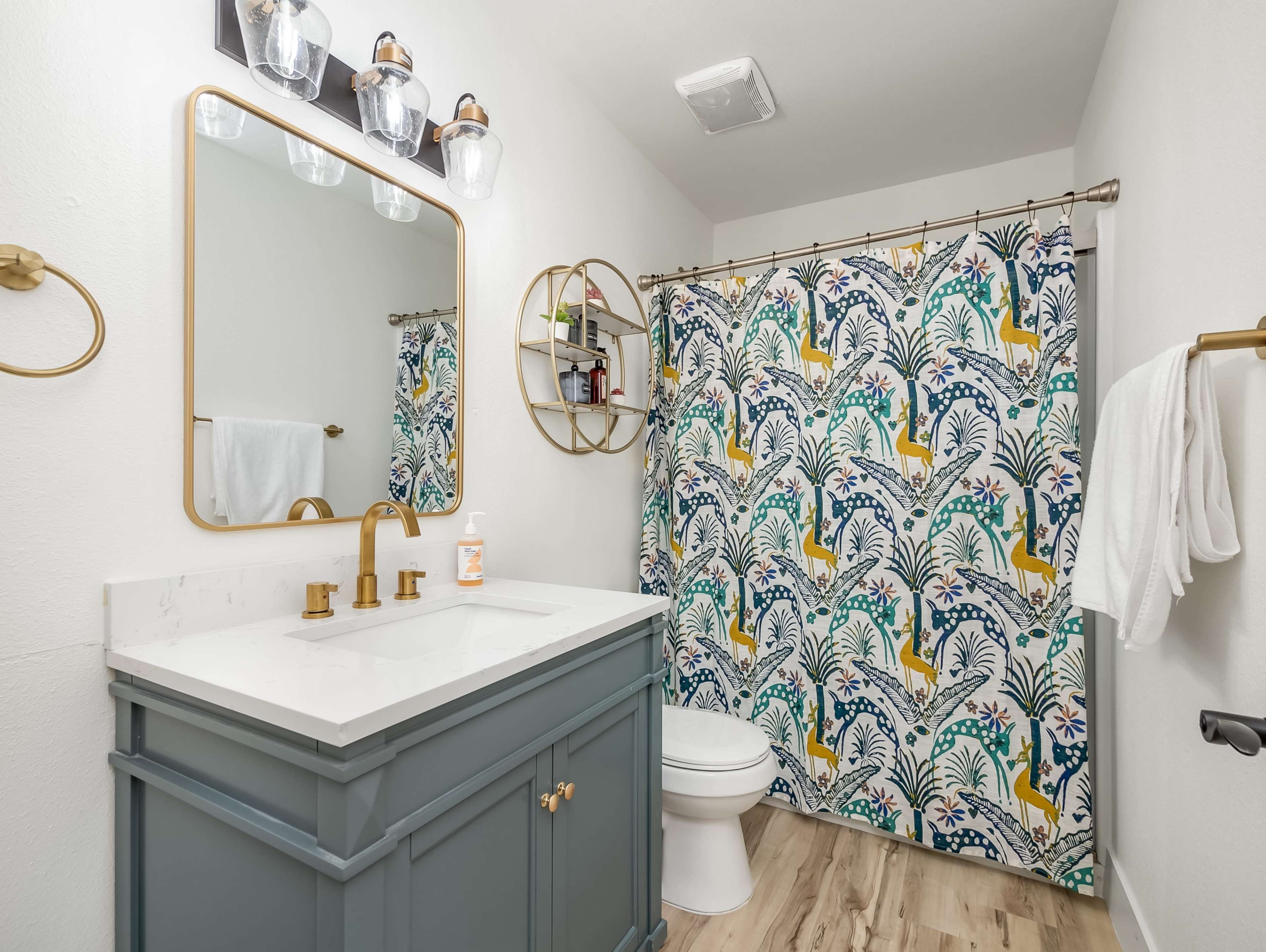 A bathroom with a blue vanity, gold fixtures, a round mirror, and a colorful patterned shower curtain.