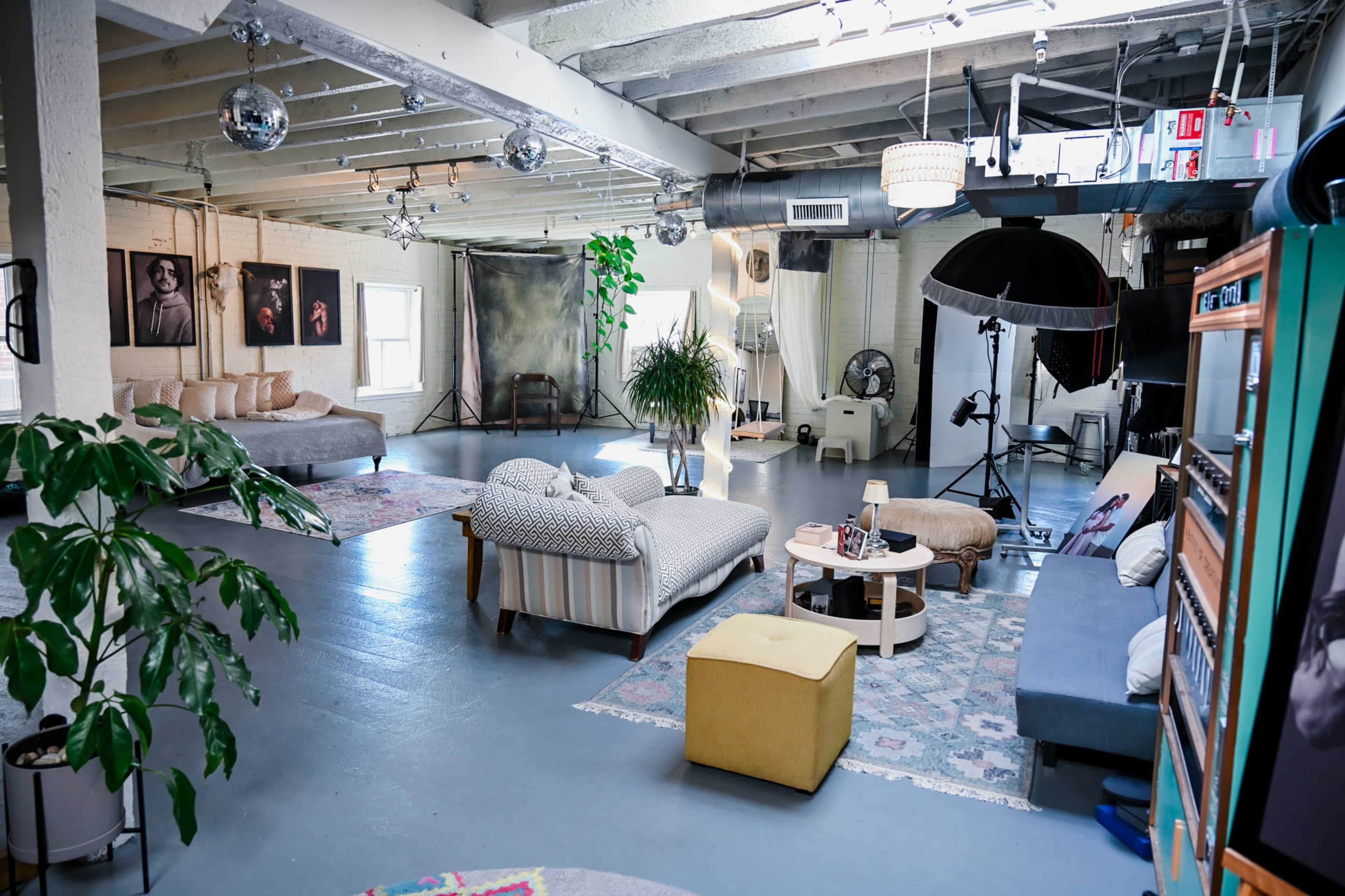 The image shows a spacious studio with various seating arrangements, a large window, and several photography lights set up for a shoot.