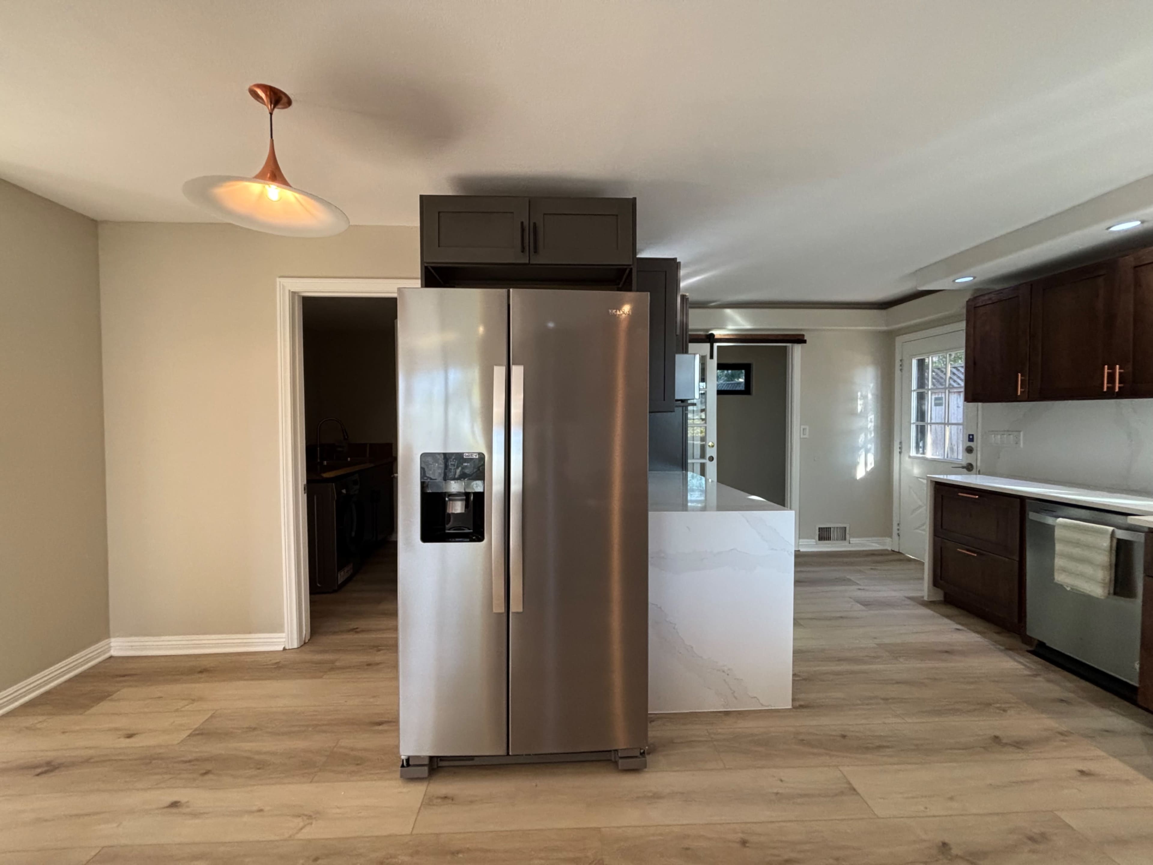 The image shows a modern kitchen featuring a stainless steel refrigerator, a white marble countertop, and wooden cabinets against a light-colored wall.
