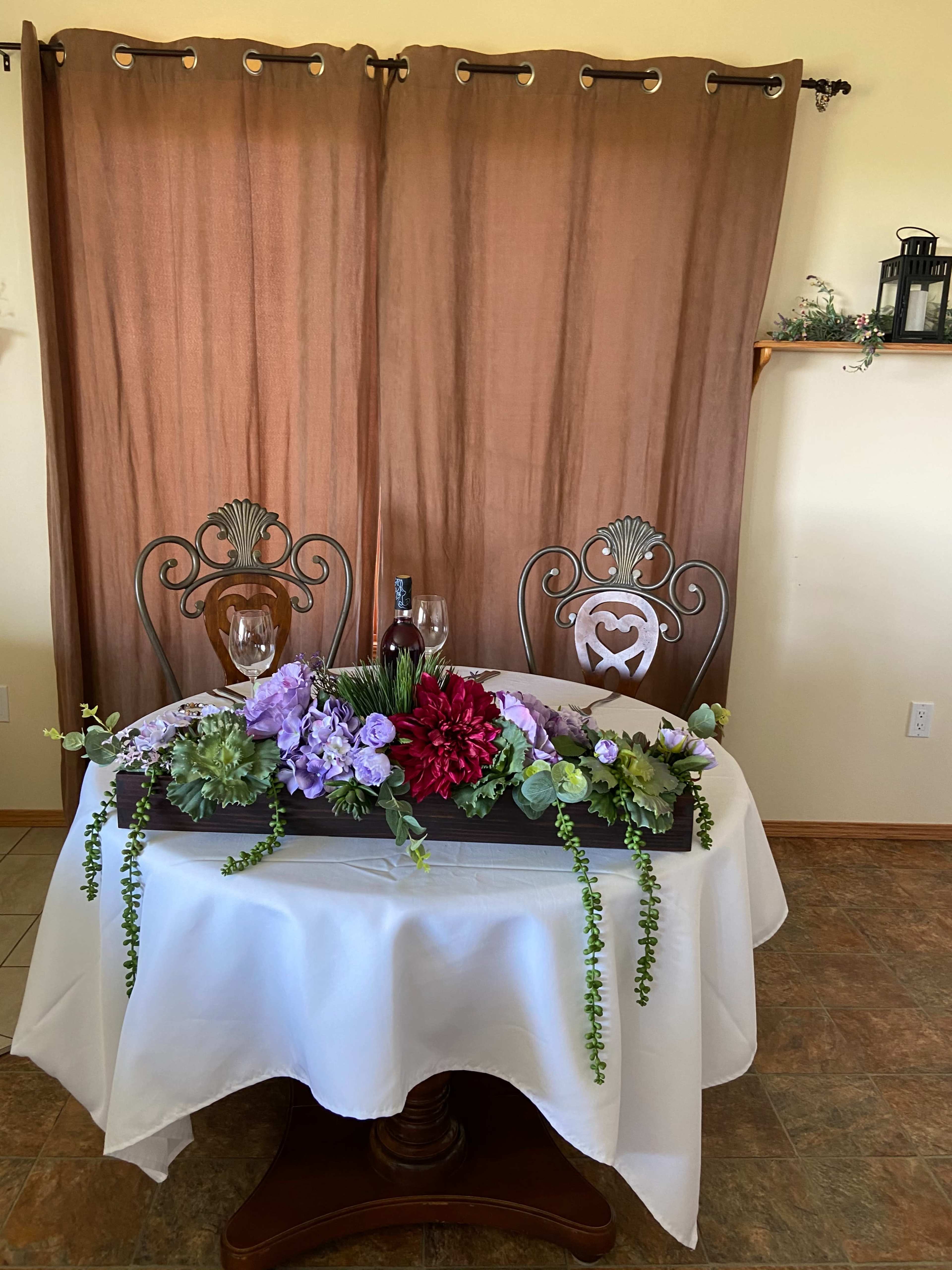 A table set for two is decorated with a floral arrangement and two glasses, positioned against a backdrop of brown curtains.