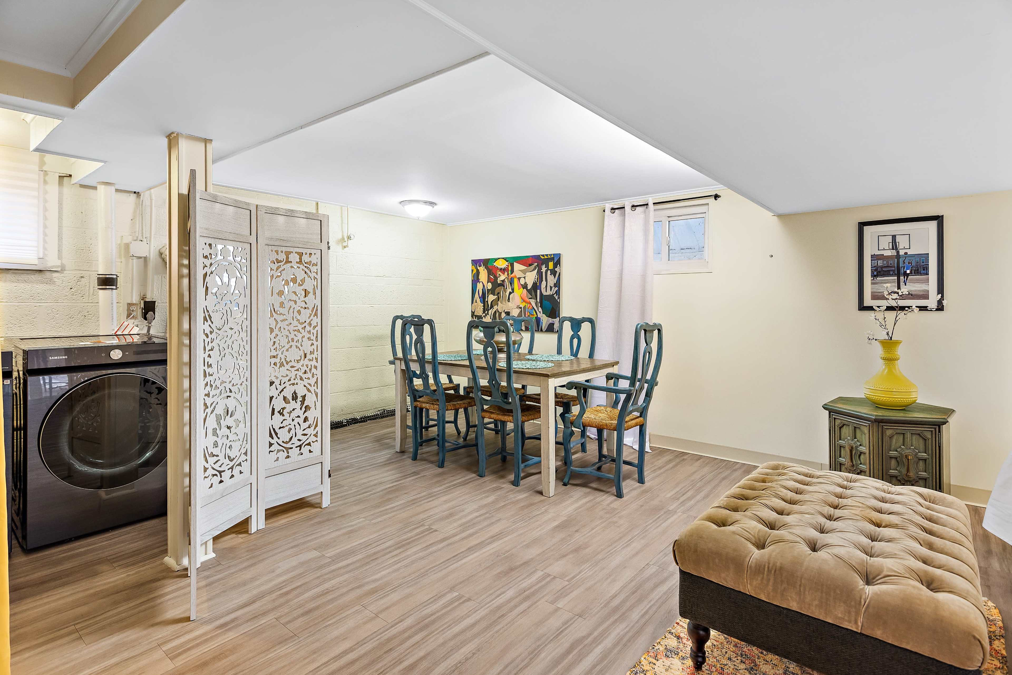 A basement area featuring a laundry space with a washing machine, a dining table with chairs, and decorative elements like a partition and artwork on the walls.