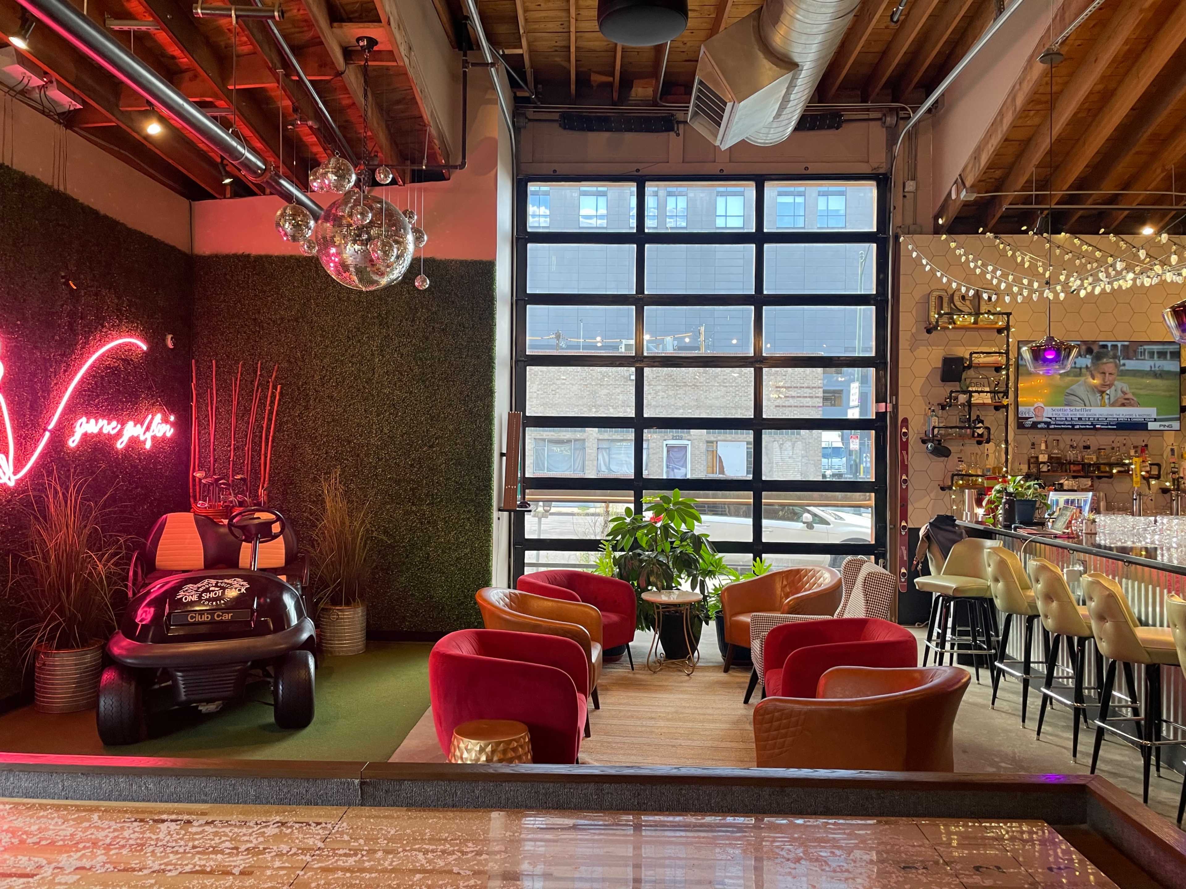A lively indoor lounge area featuring a bar, colorful seating, a golf cart decoration, and a large glass door opening to an outdoor view.