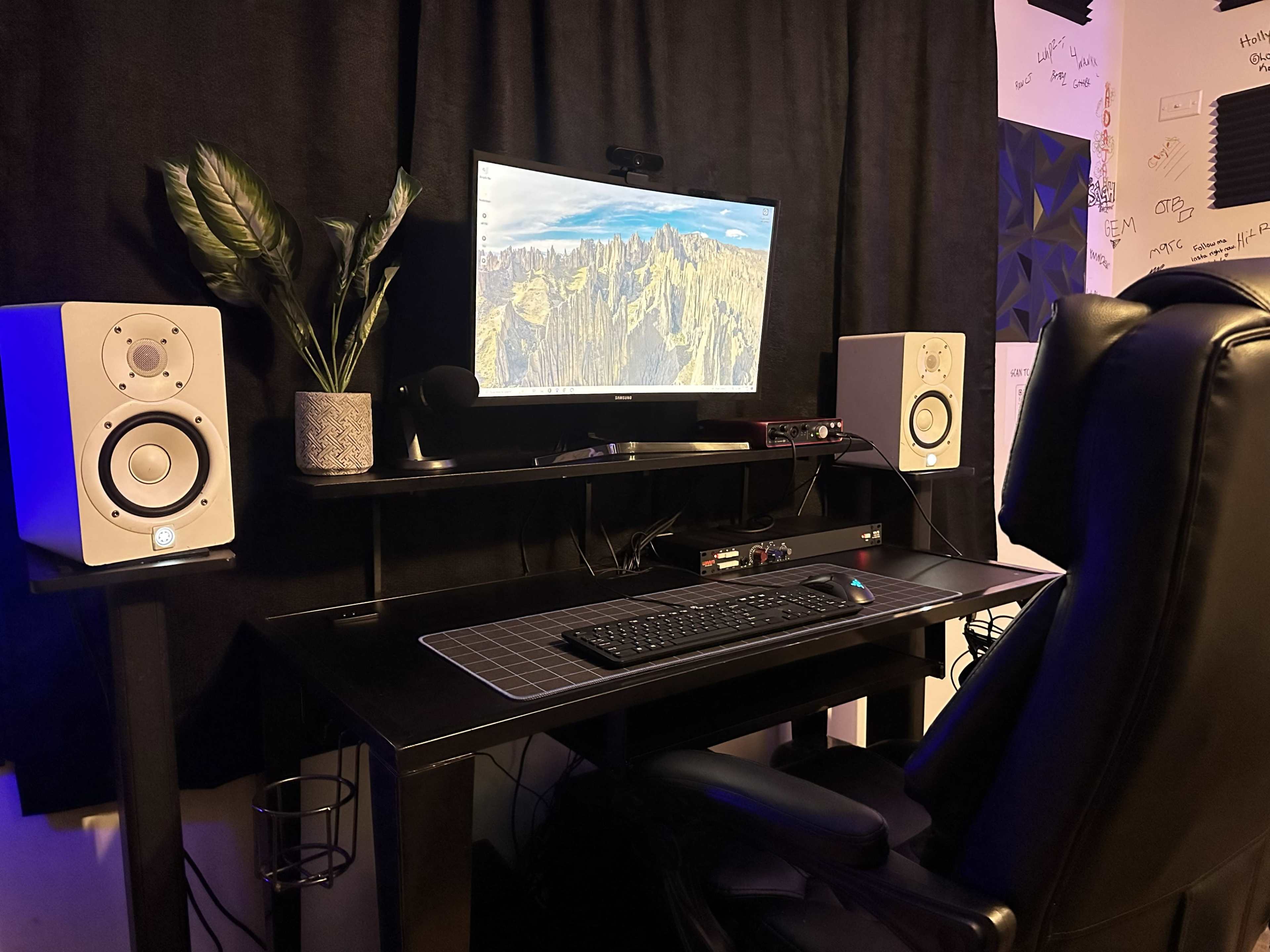 A modern workspace features a large monitor on a black desk, flanked by speakers, with a keyboard and mouse in front and a plant nearby.