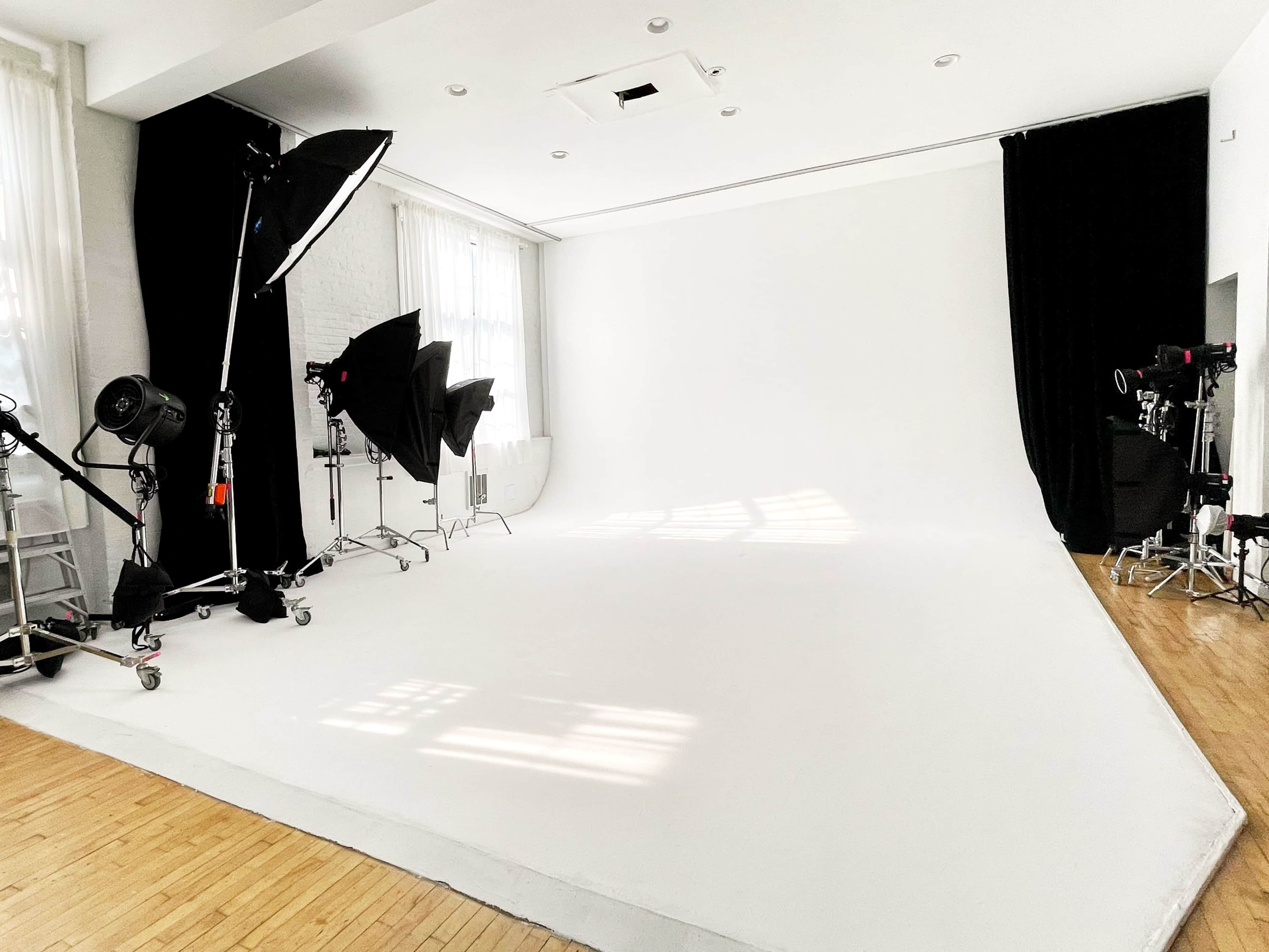 A photography studio with a large white backdrop, various lighting equipment, and dark curtains.