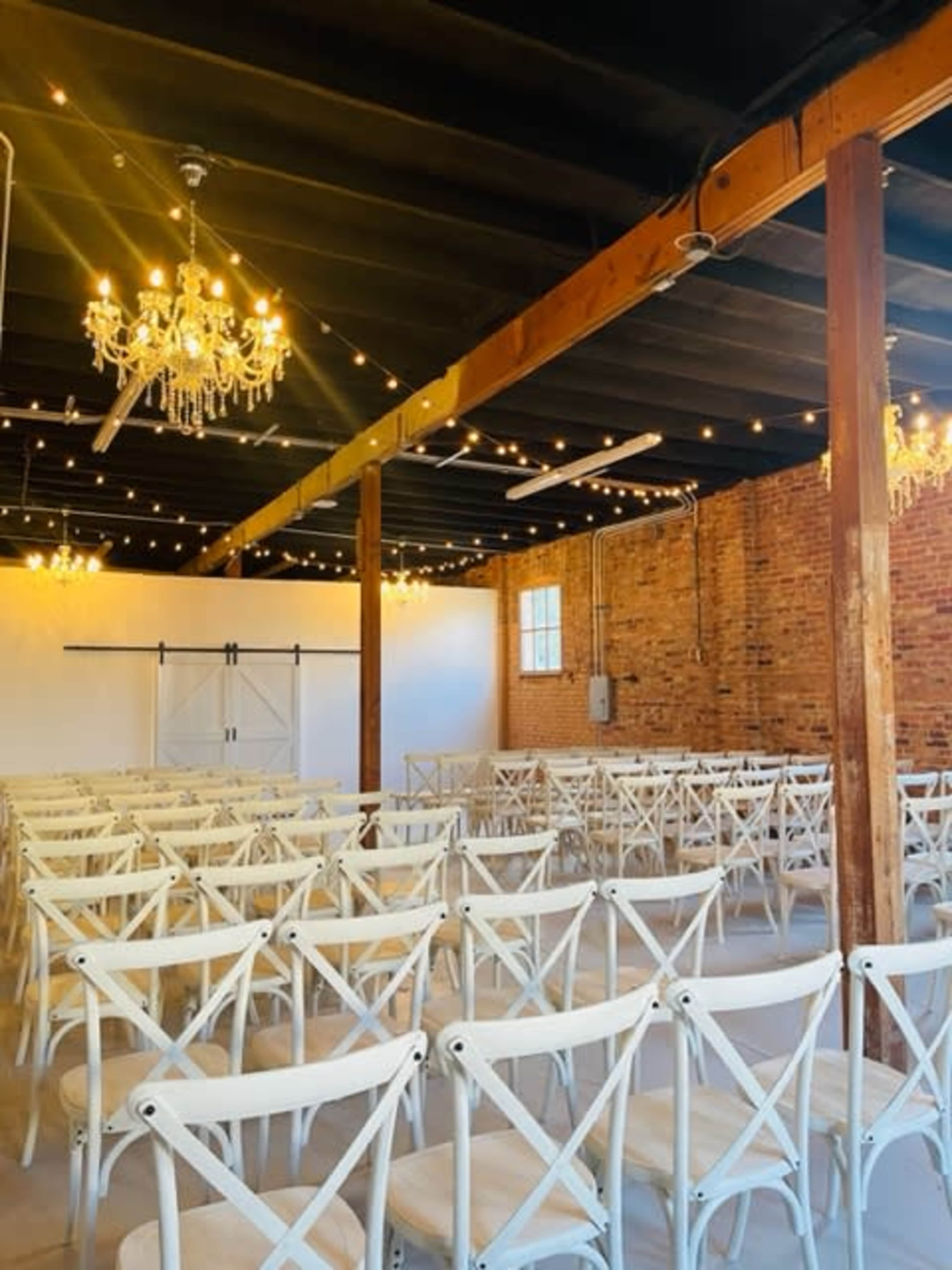 An empty event space with rows of white chairs, exposed brick walls, and hanging chandeliers illuminated by string lights.