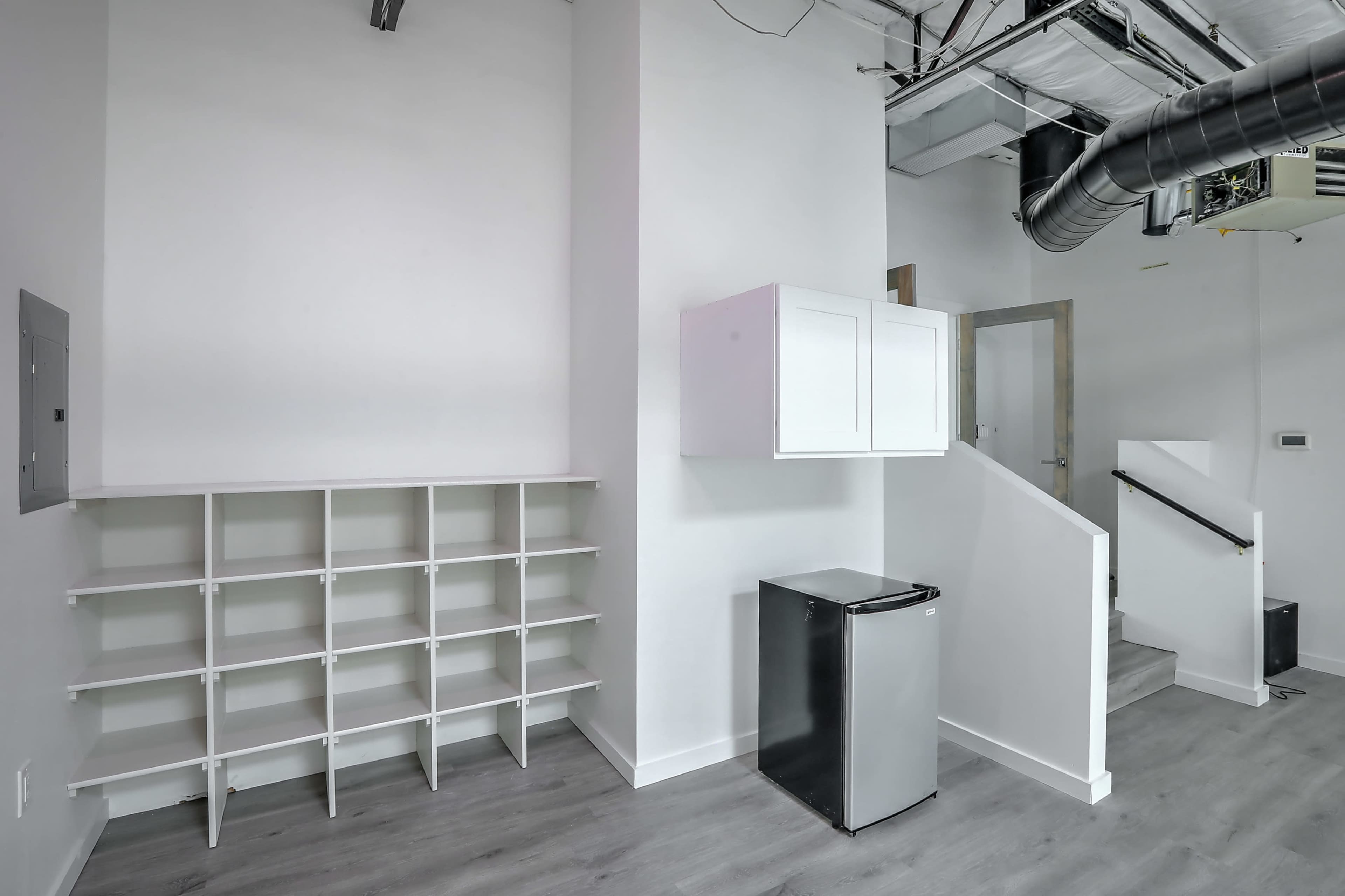The image shows a small, modern room featuring a white shelving unit, a refrigerator, and a staircase.