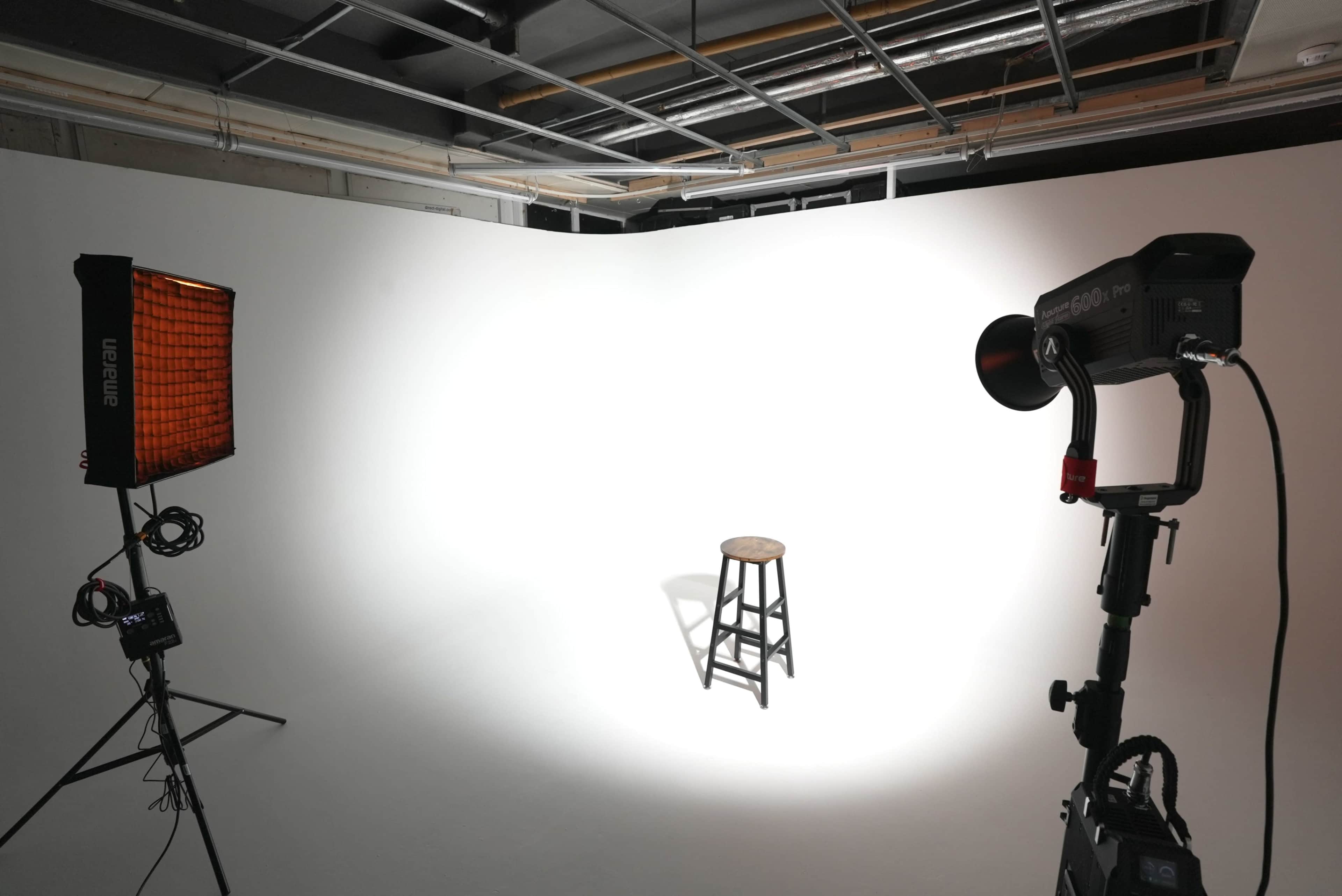 A wooden stool is positioned under a spotlight in a minimalist photography studio with a plain white backdrop.