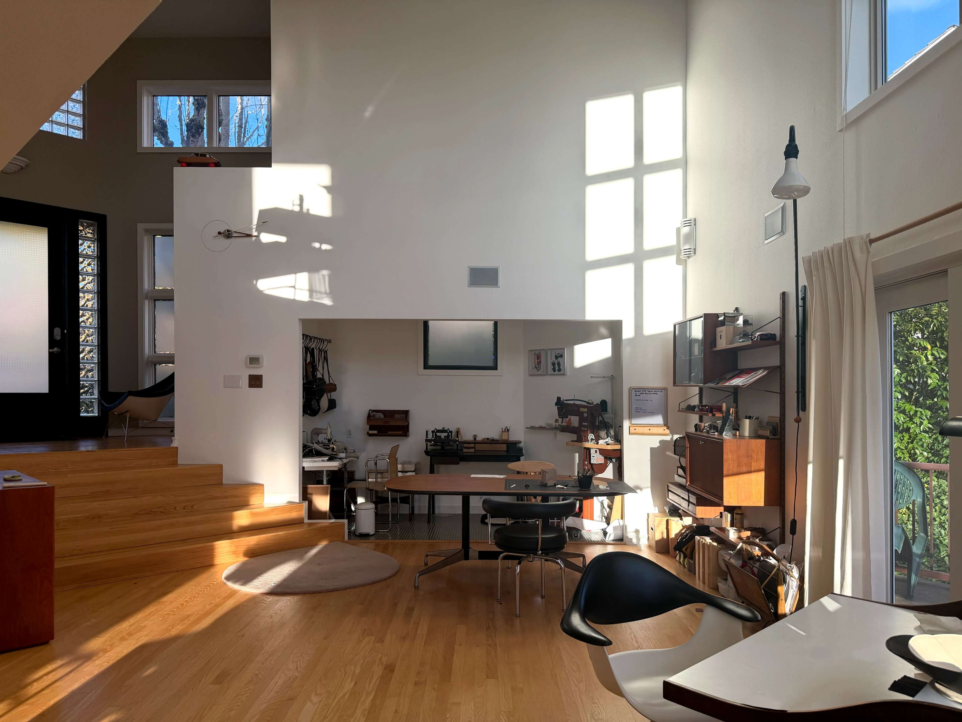 The image shows a modern, open-concept living space with wooden flooring, large windows allowing natural light, and a well-organized work area featuring a desk and various tools.