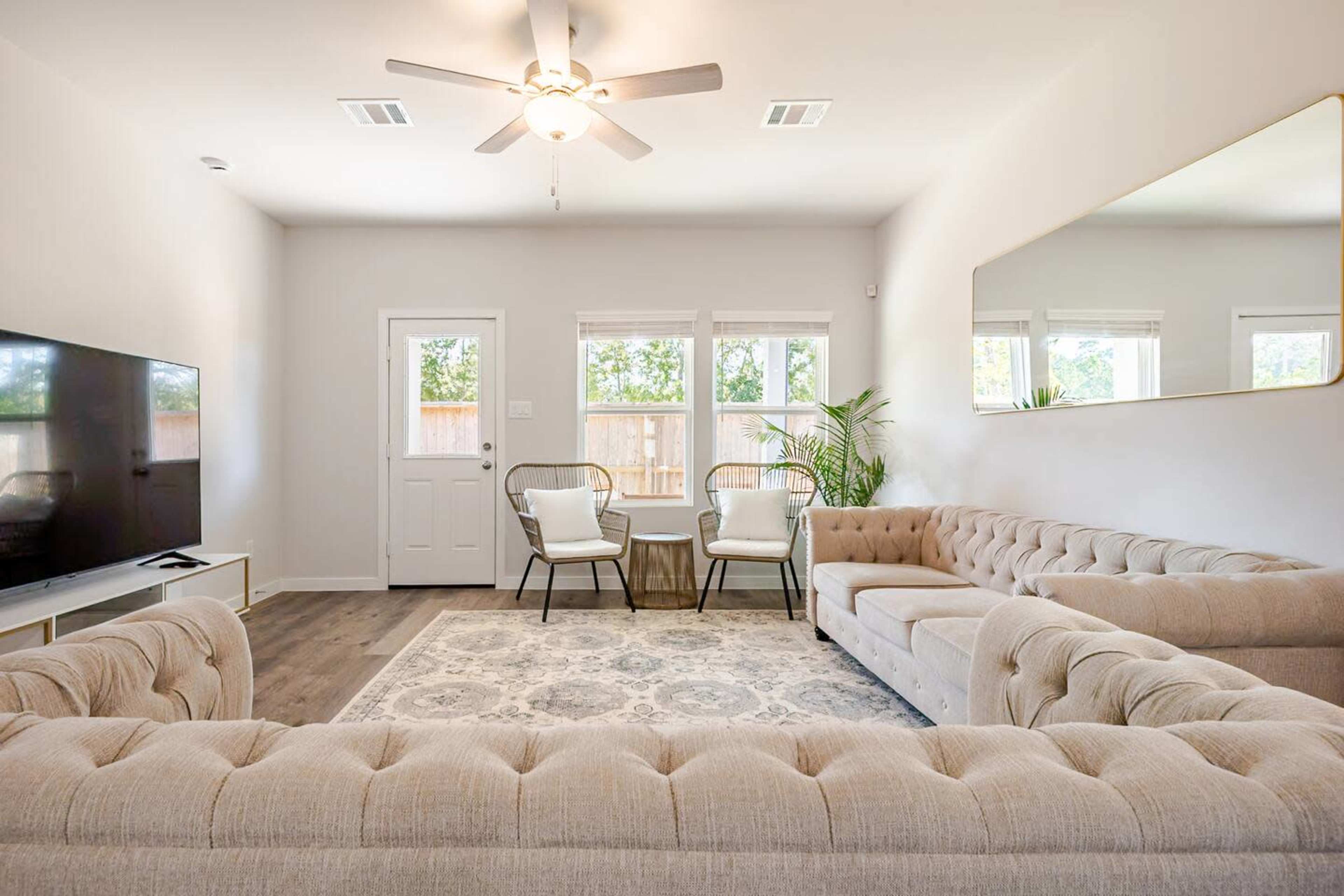 The image shows a modern living room featuring a beige tufted sectional sofa, a television mounted on the wall, two chairs near a small table, and large windows allowing natural light.