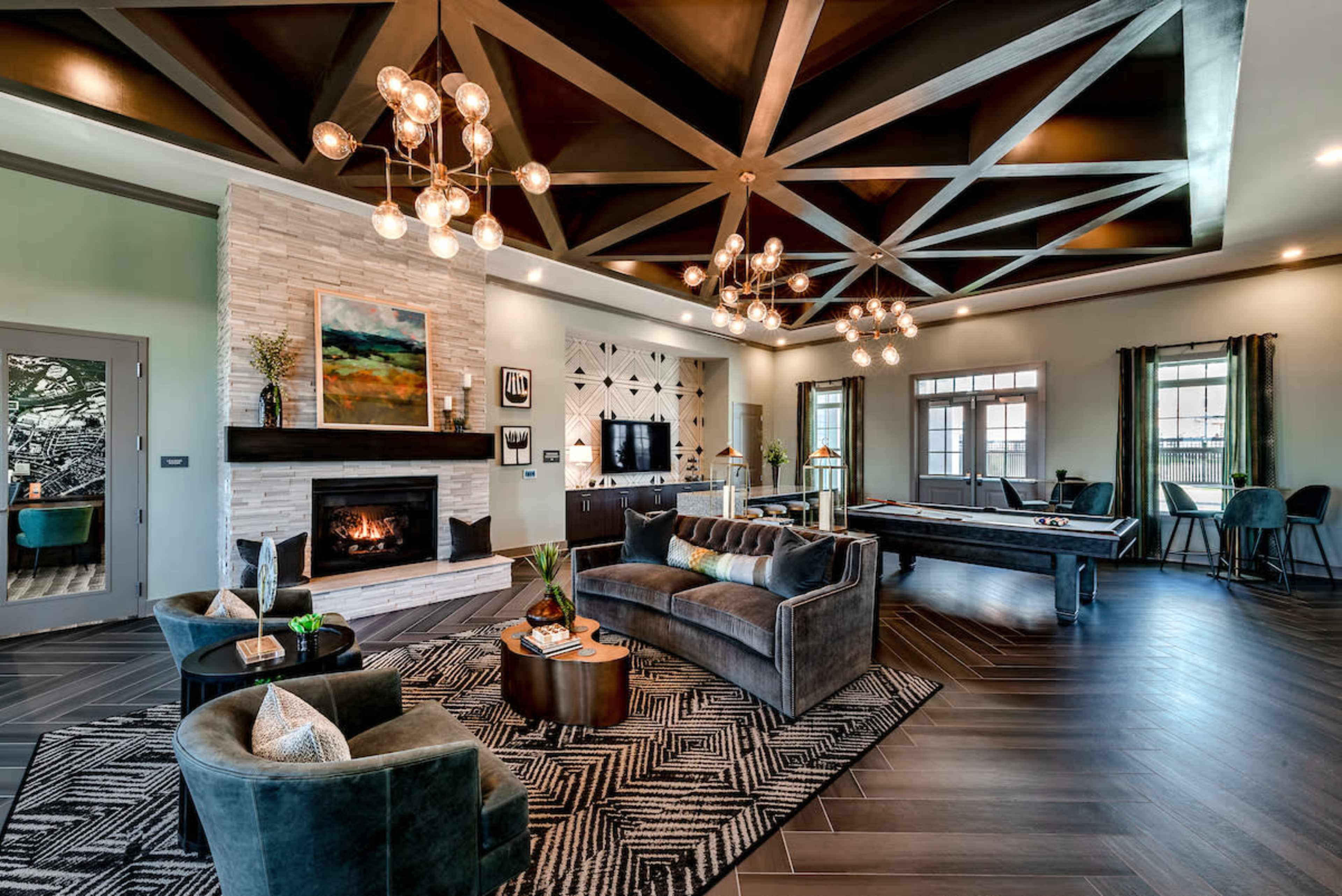 The image shows a modern living room featuring a stone fireplace, dark wood accents, large windows, and a pool table, all illuminated by decorative overhead lighting.