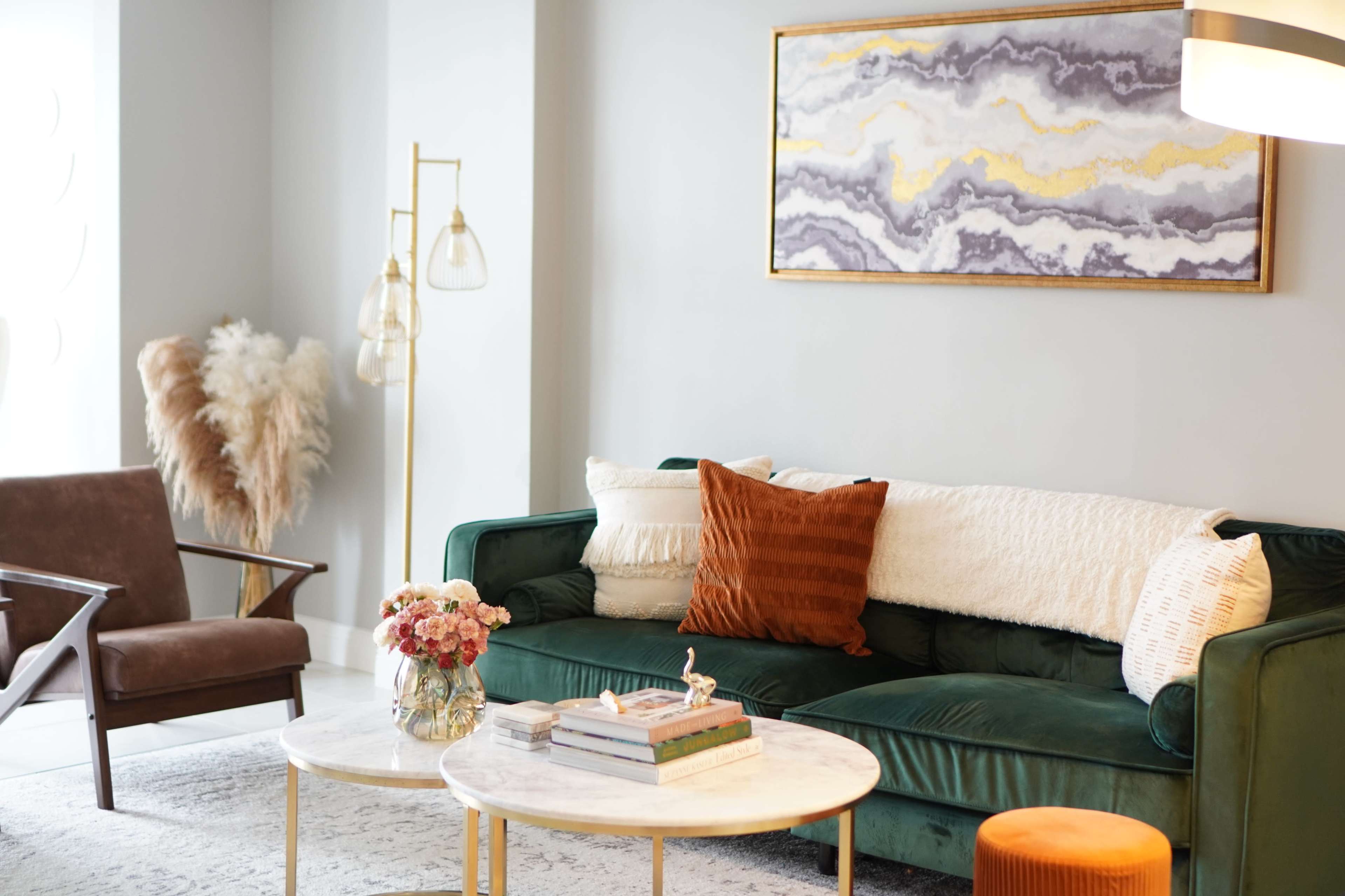 A modern living room features a green velvet sofa with decorative pillows, a marble coffee table, and a stylish artwork above, complemented by a floor lamp and floral arrangements.