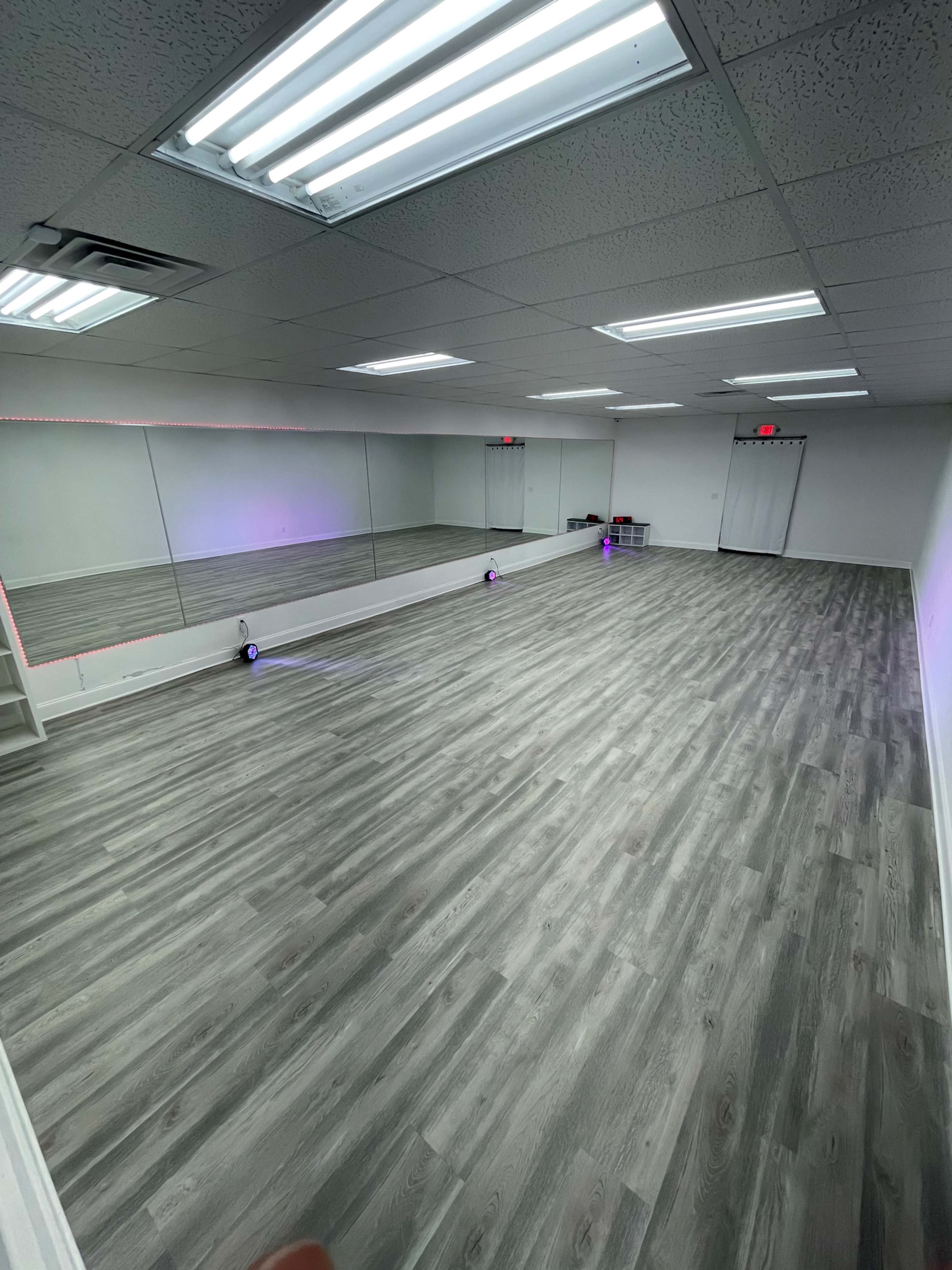 An empty studio space with gray hardwood flooring, a large mirror along one wall, and bright overhead lighting.