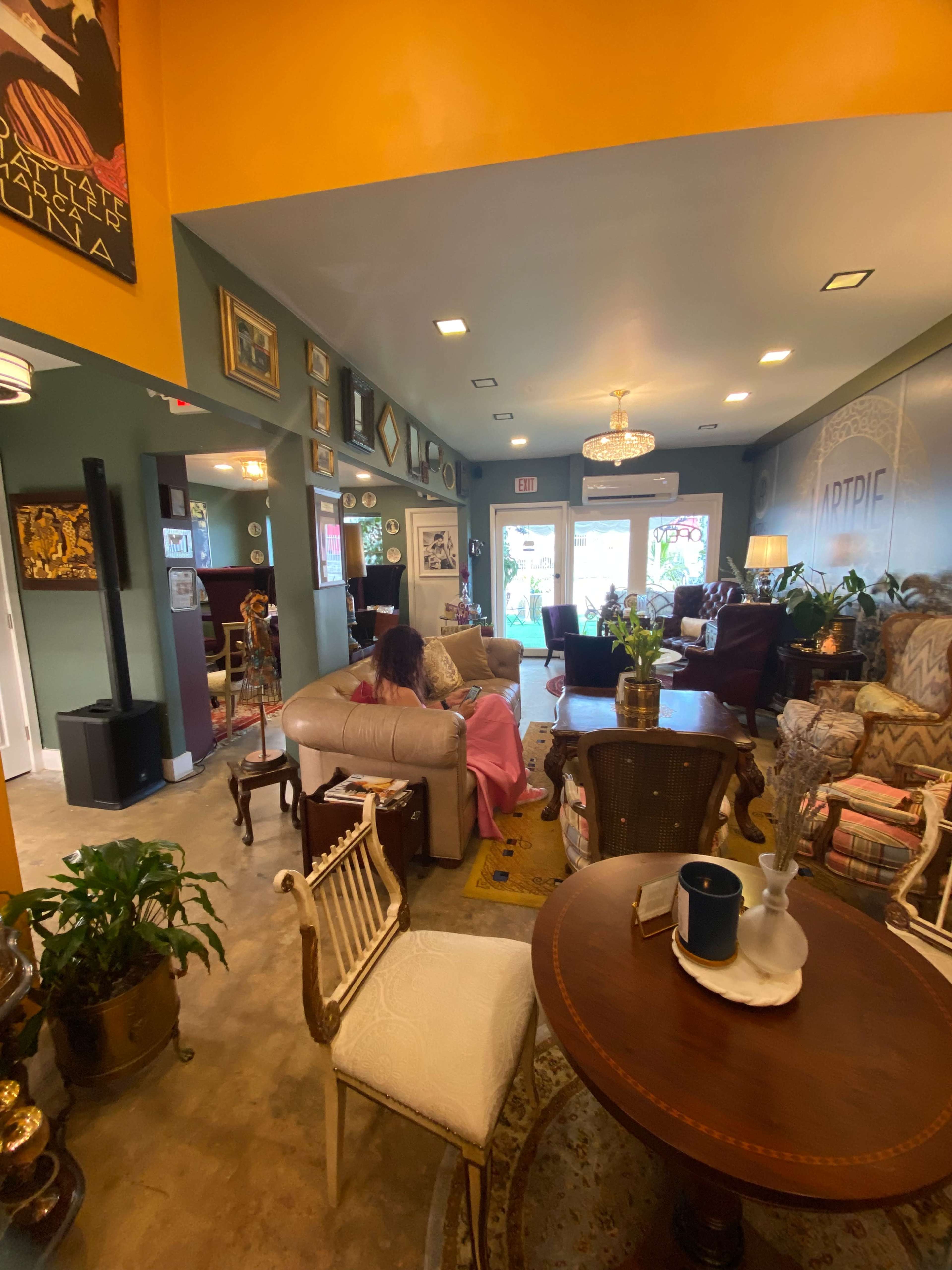 A cozy, eclectic interior of a café or lounge with various seating areas, decorative art on the walls, and large windows letting in natural light.