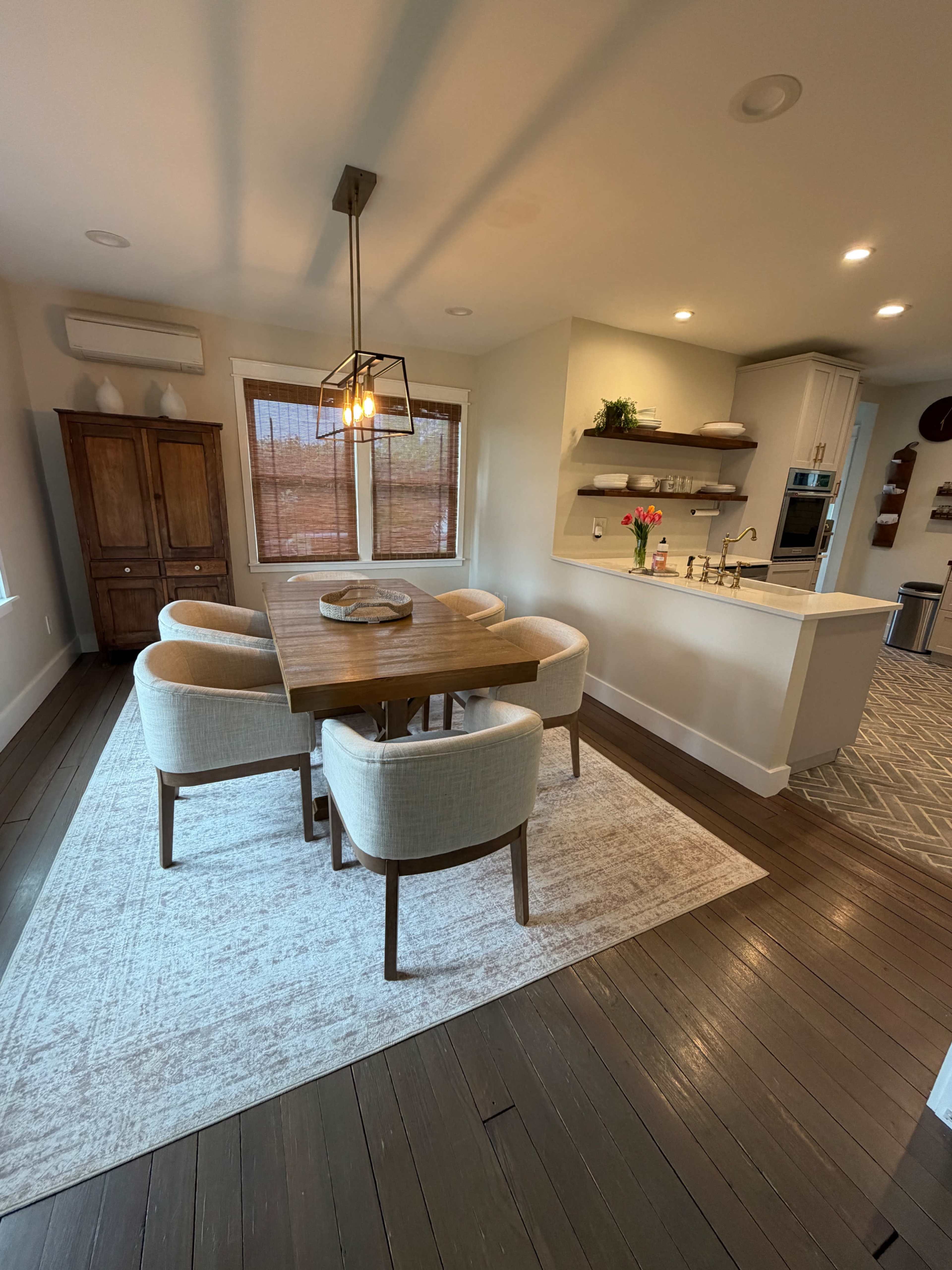 The image shows a modern dining area with a wooden table surrounded by four upholstered chairs, a light fixture above the table, and a wall-mounted shelf displaying decor.