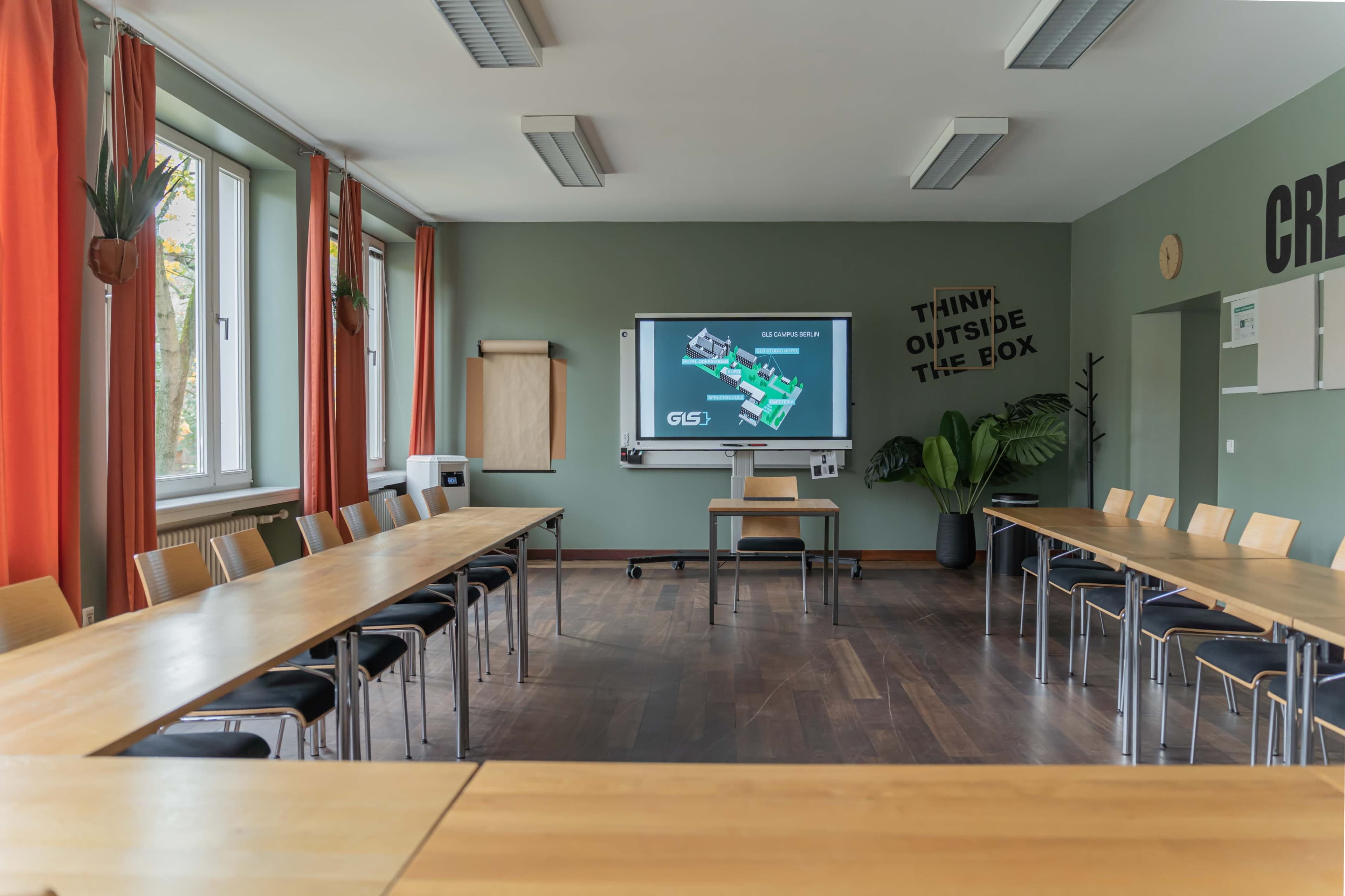 A conference room with a large table arranged in a U-shape, featuring a projector screen displaying a presentation, surrounded by wooden chairs and plants.