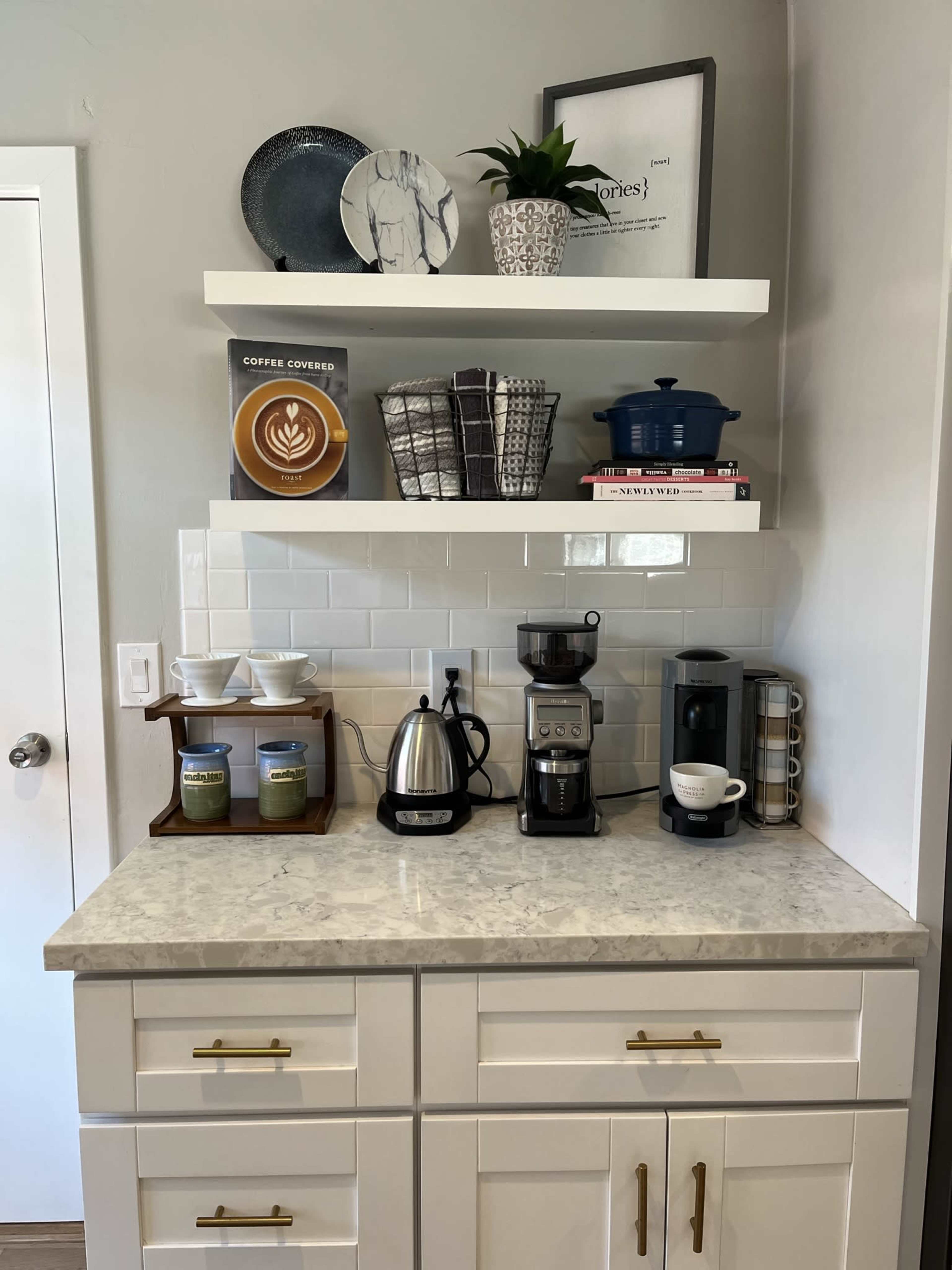 A kitchen countertop with a coffee maker, electric kettle, and a collection of cookbooks and decorative items on display shelves above.