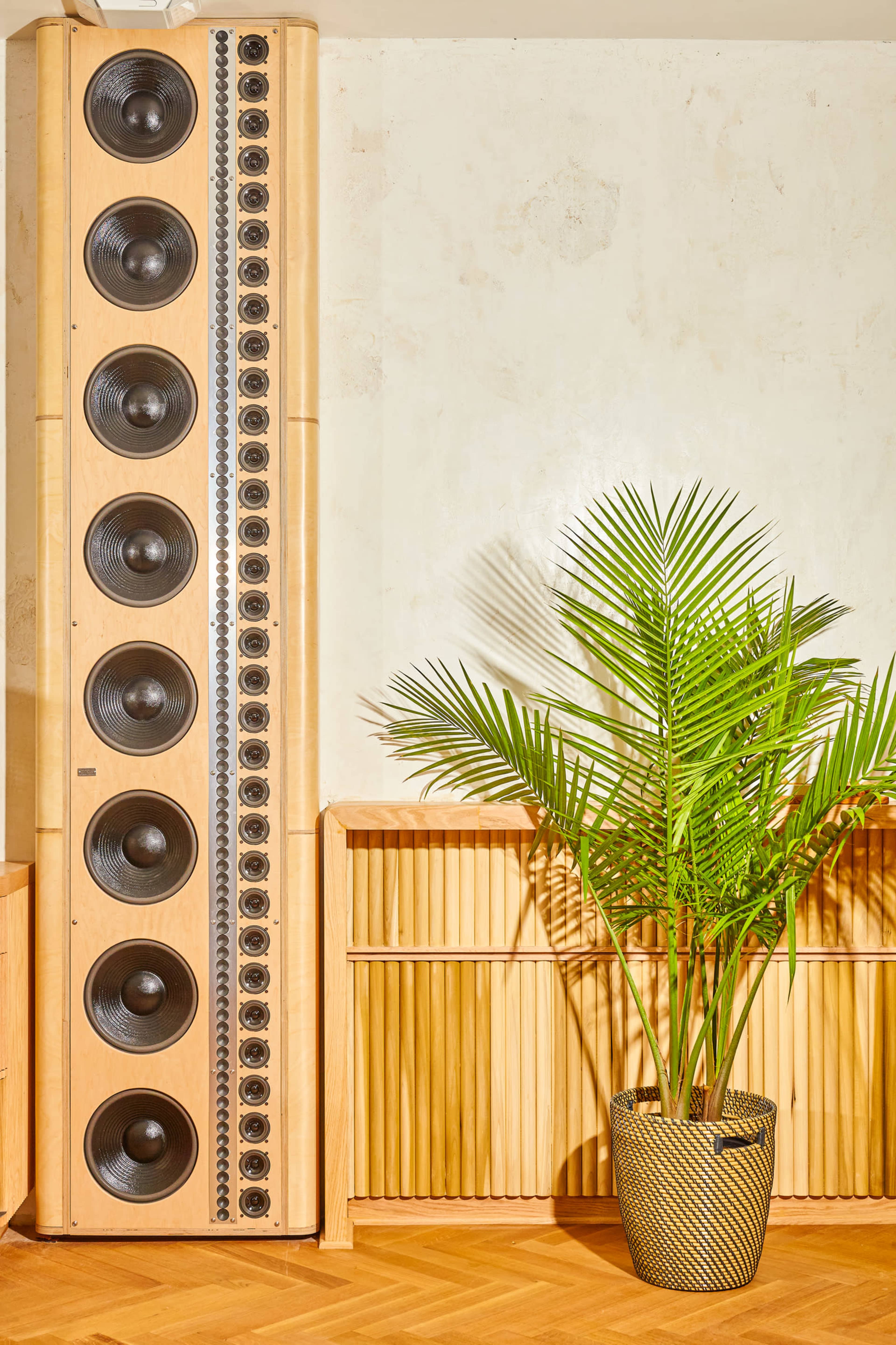 A tall speaker with multiple woofers stands next to a potted palm plant in a room with a textured wall.