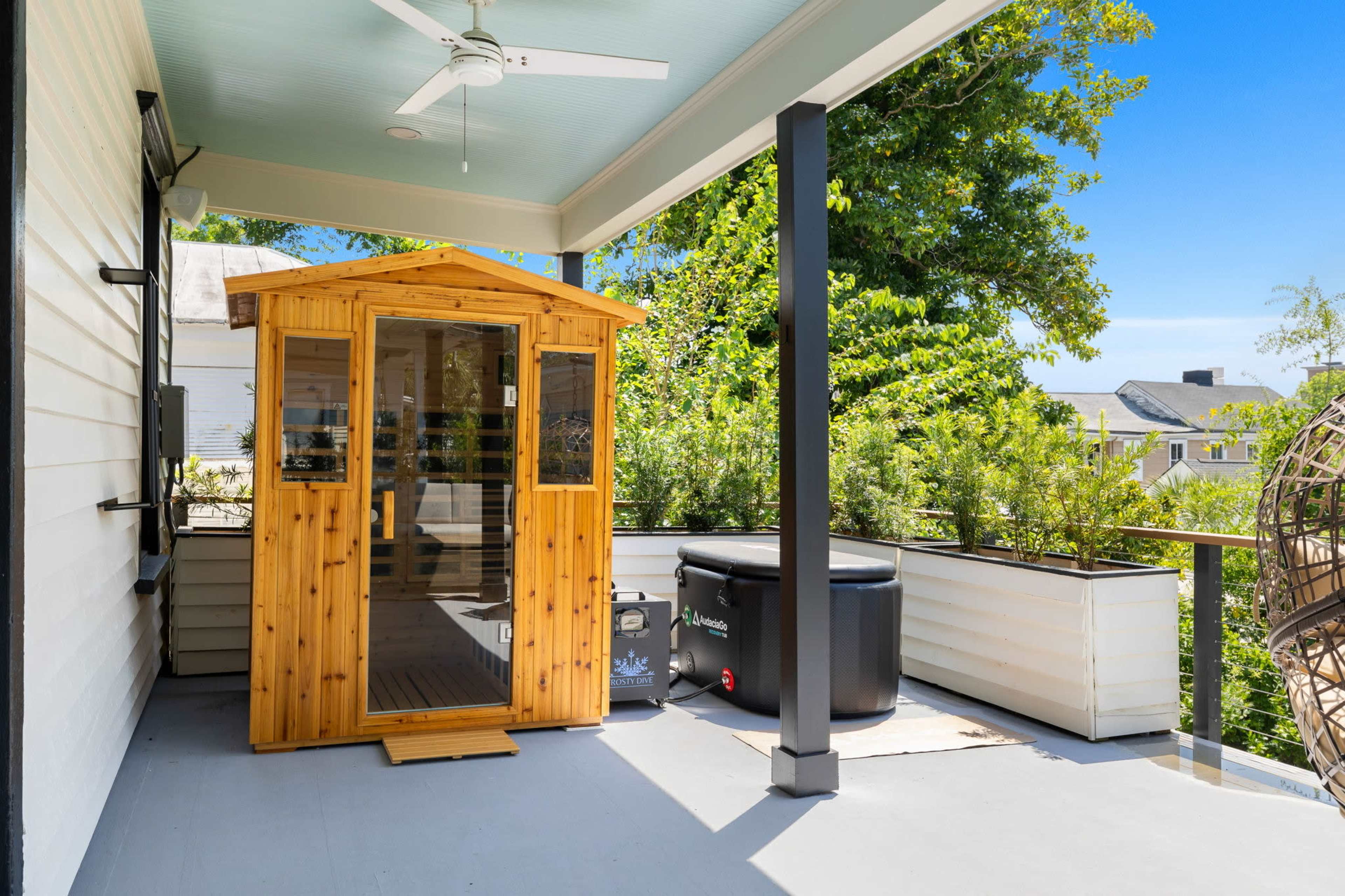A wooden sauna with large glass doors is situated on a porch beside a black hot tub, surrounded by greenery and clear blue skies.