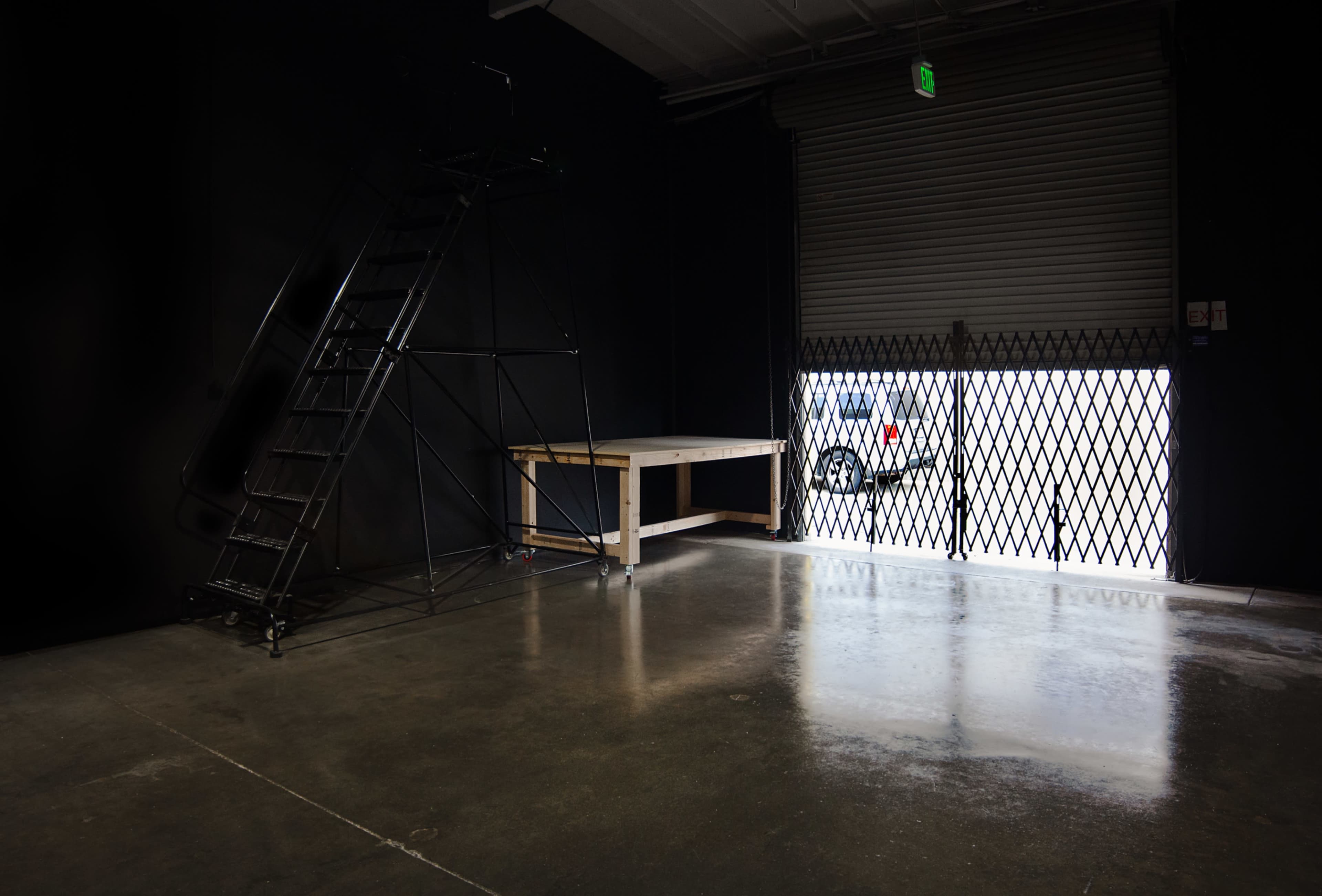 The image shows a dimly lit industrial space with a metal staircase on the left and a secured garage door on the right, revealing a parked vehicle outside.