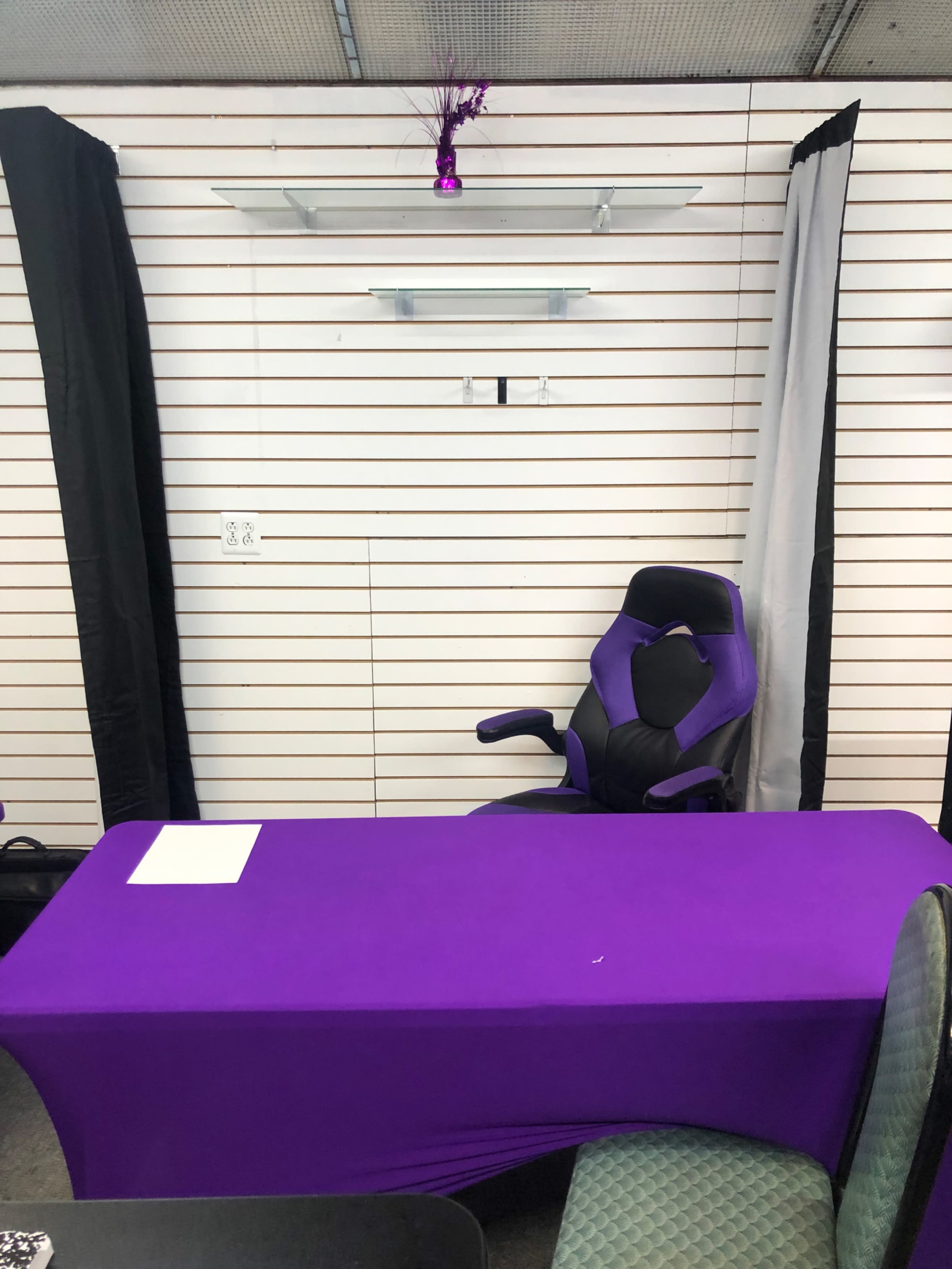 A purple table is situated in front of a black and white backdrop with a shelf above it and an ergonomic chair beside the table.