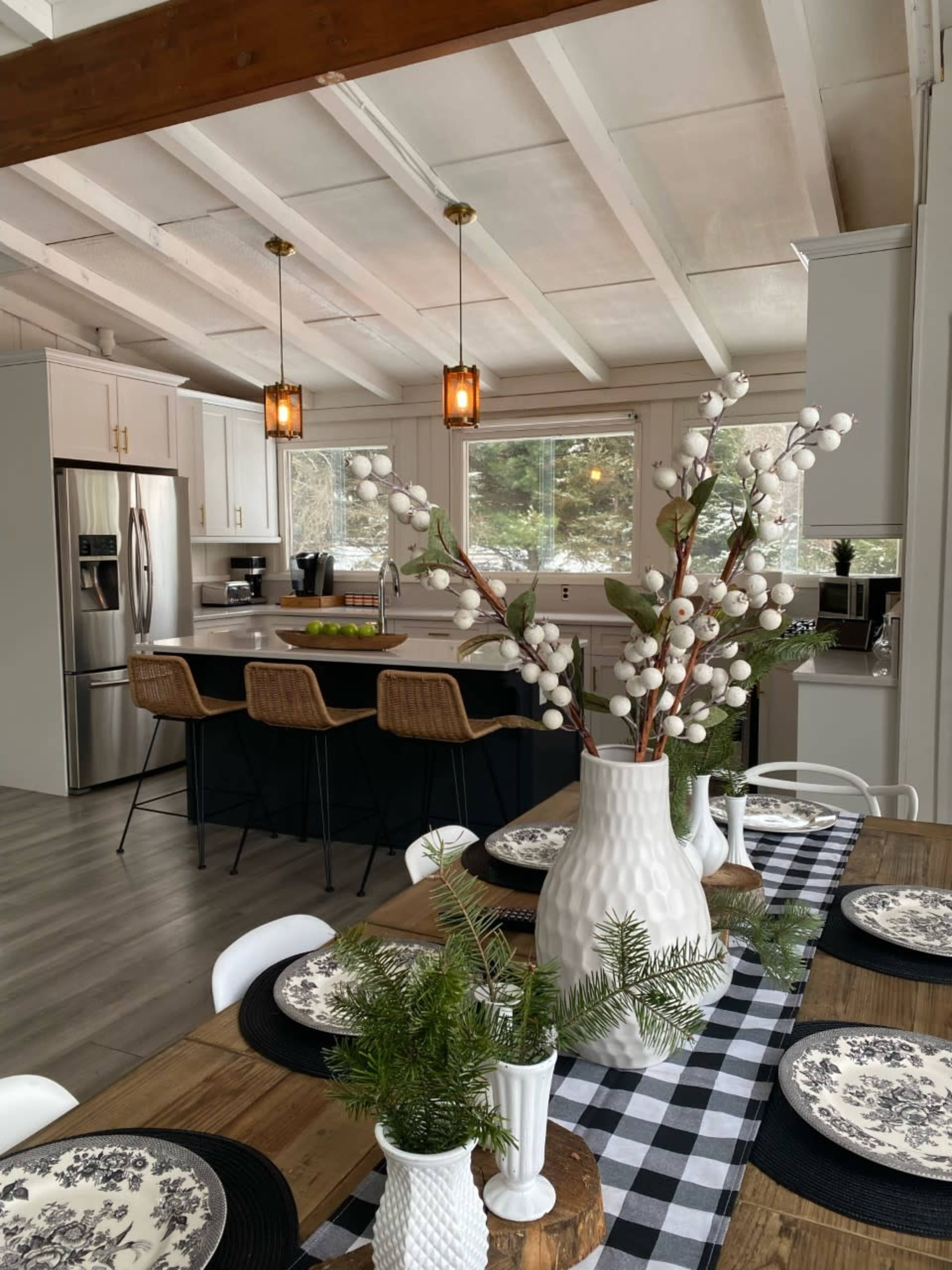 A modern kitchen and dining area featuring a wooden table set with black and white patterned plates, a decorative vase with cotton branches, and a kitchen island with bar stools.