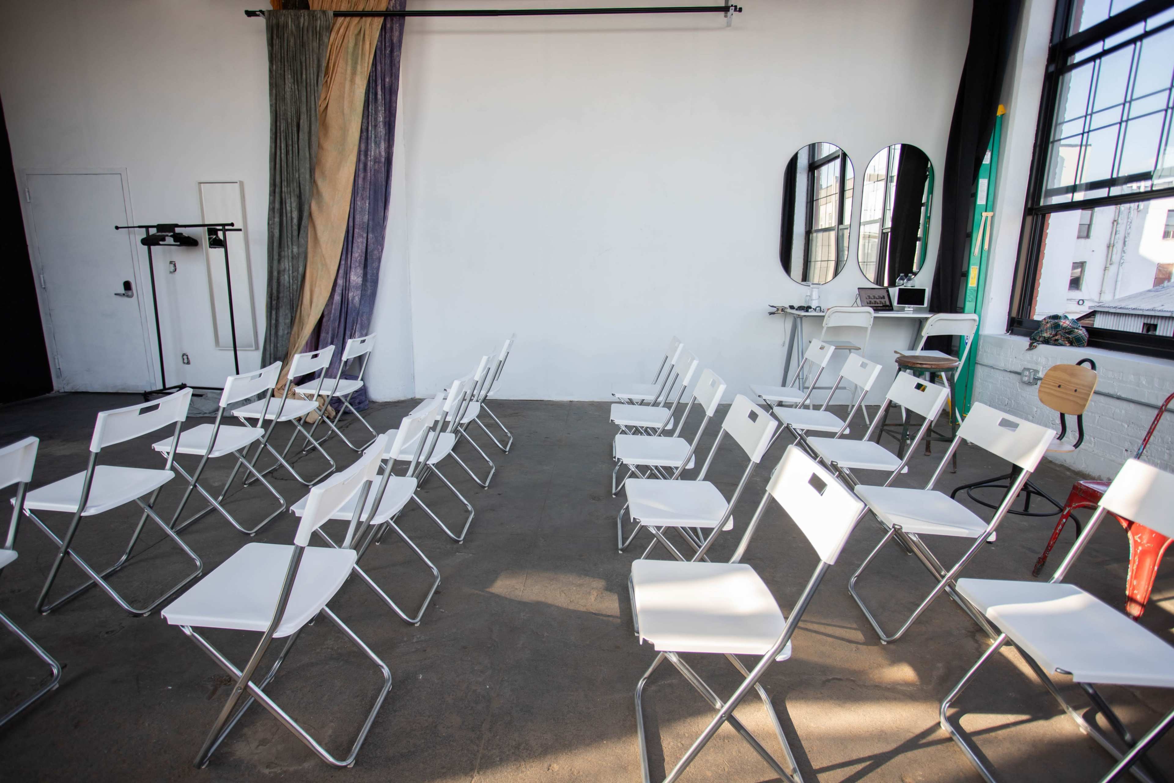 A spacious room with white walls arranged with several rows of white folding chairs and a large mirror setup against the backdrop.