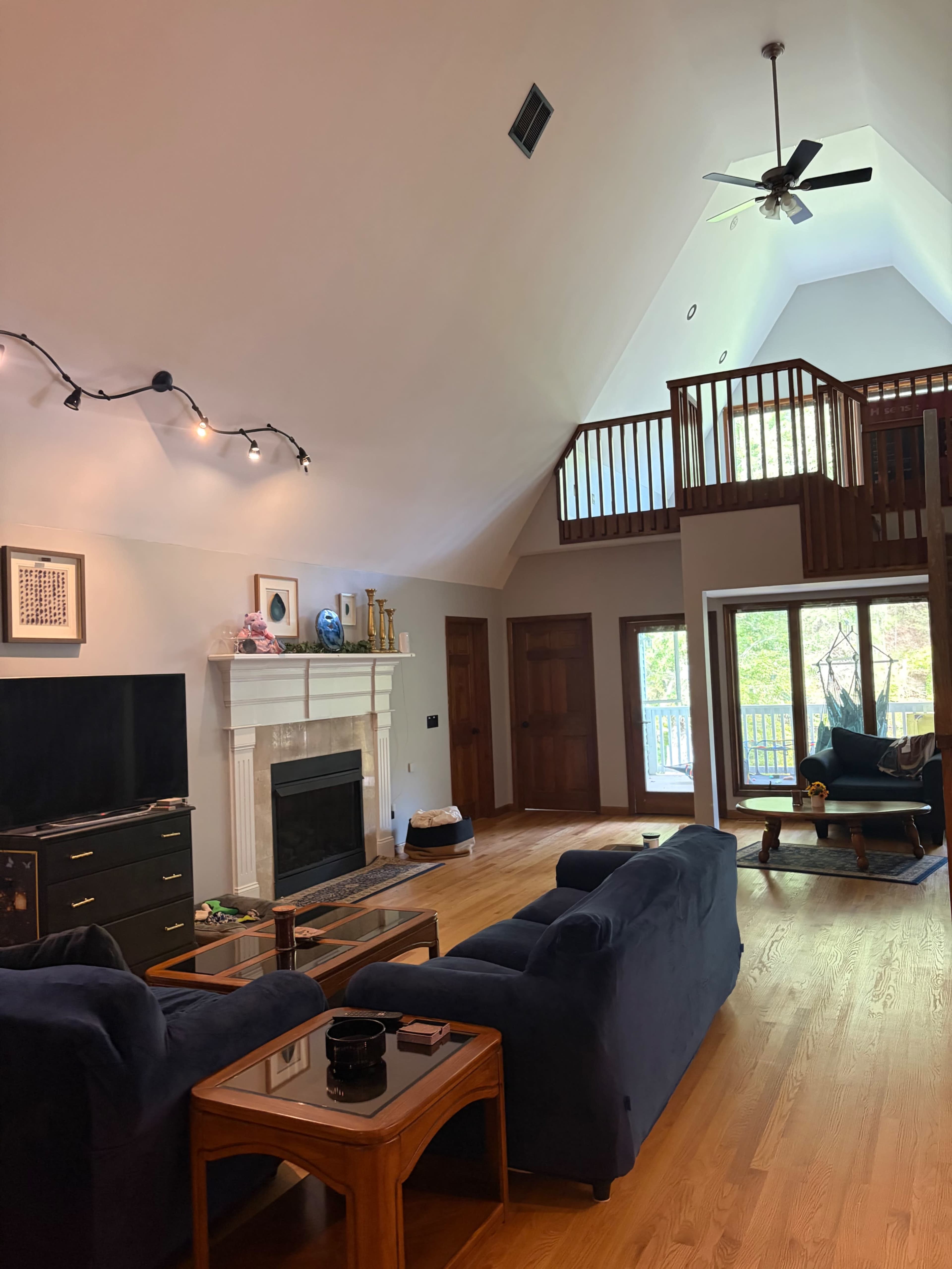 The image shows a spacious living room with a high ceiling, featuring a fireplace, two dark blue sofas, a coffee table, and a large window along with a balcony above.
