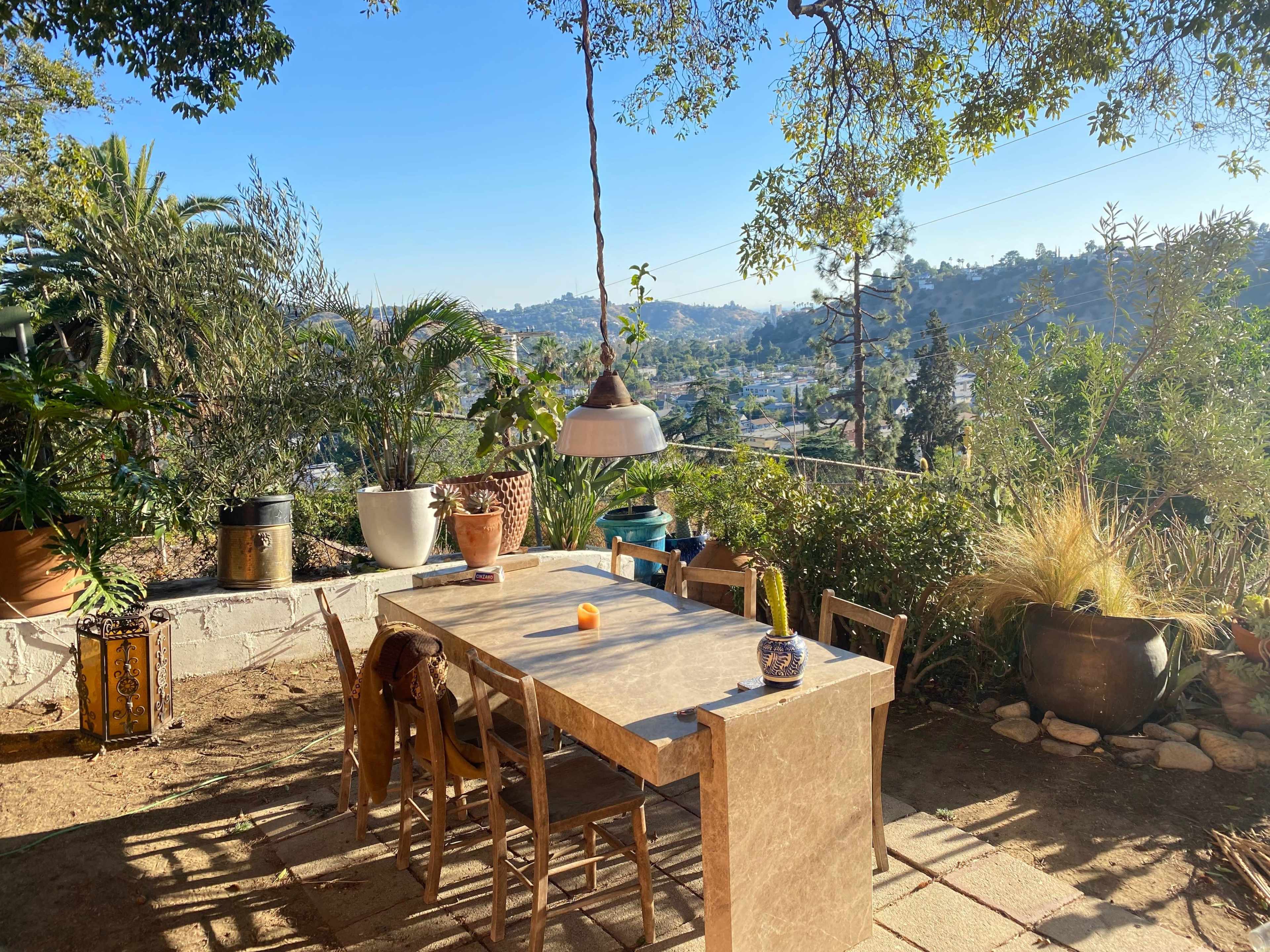 A patio table with wooden chairs is set outdoors, surrounded by potted plants and overlooking a view of hills and houses.