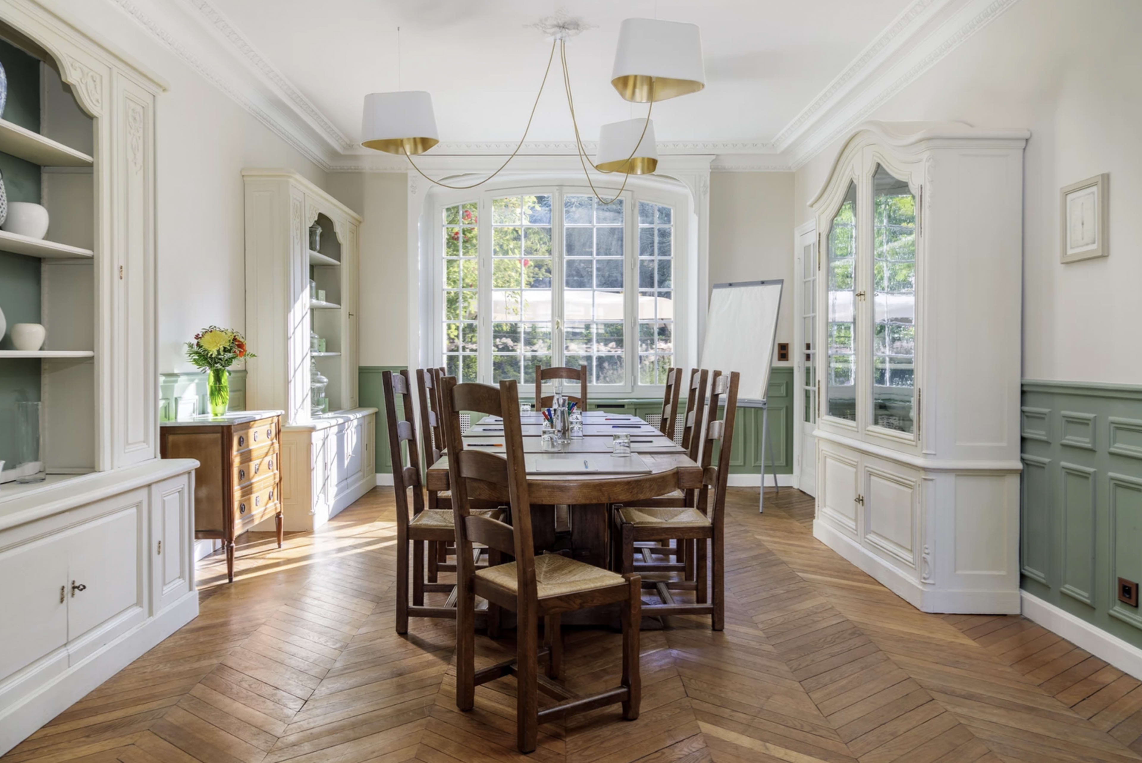 A wooden dining table surrounded by chairs is situated in a well-lit room with large windows, decorative cabinetry, and a herringbone-patterned floor.