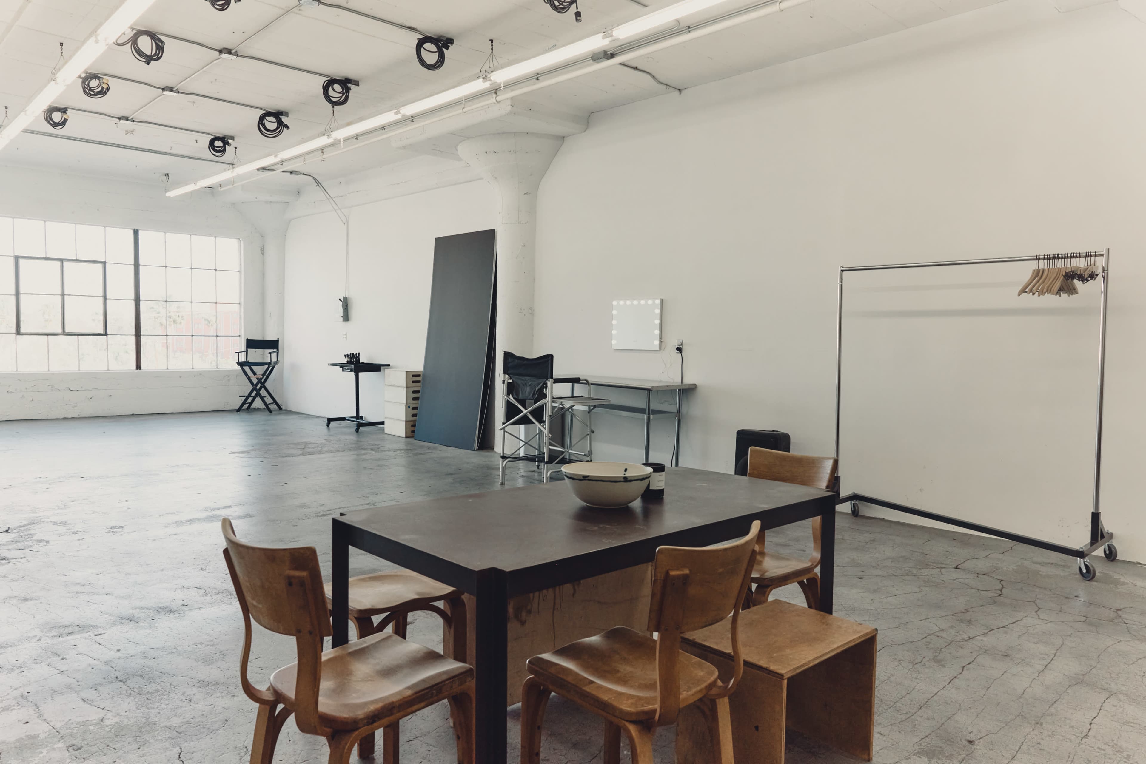 The image shows a spacious, empty studio with a large window, a wooden table surrounded by chairs, and a clothing rack positioned against the wall.