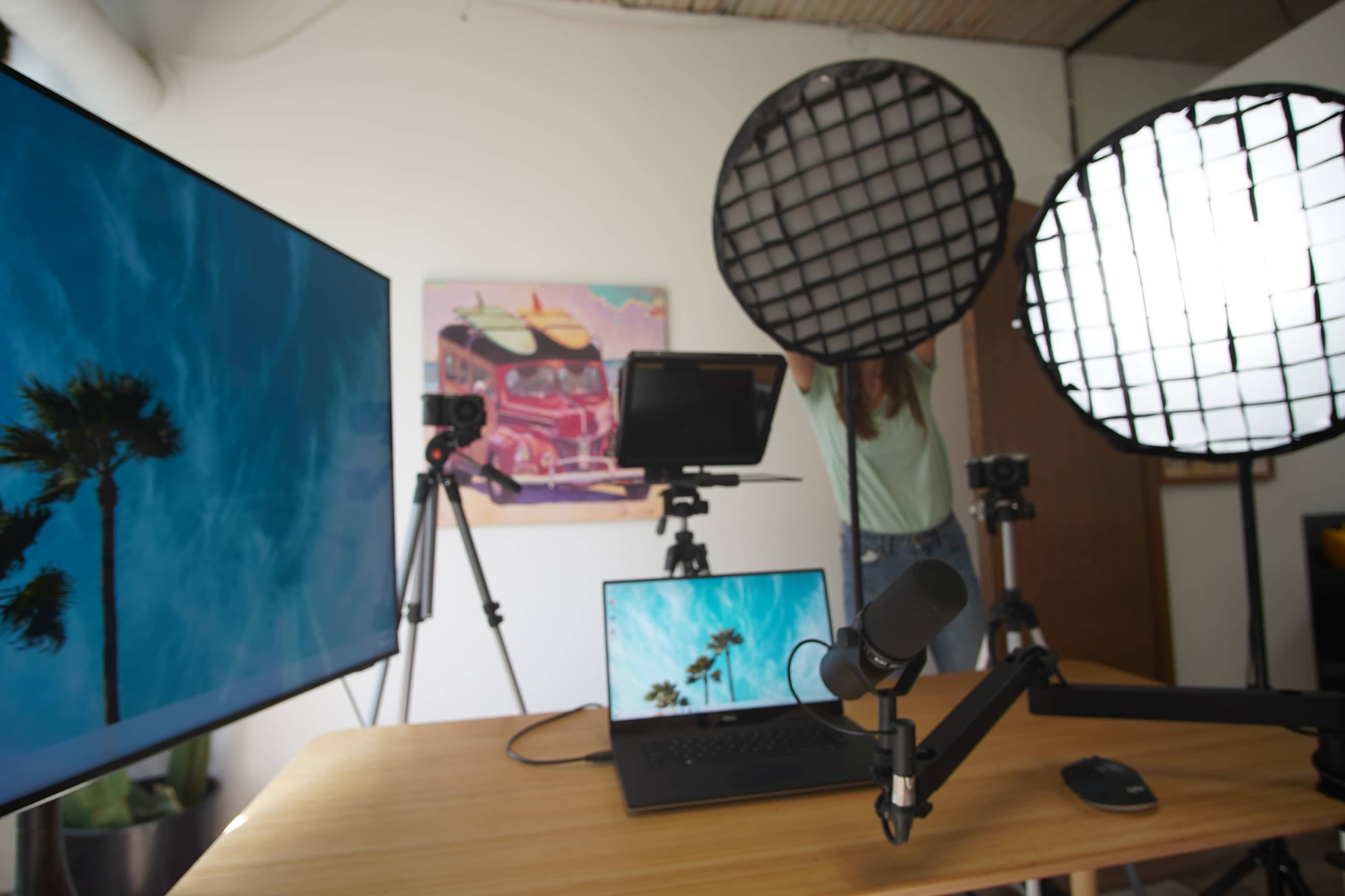 A workspace is set up with a laptop, a monitor displaying a palm tree, and various cameras and lights arranged on tripods.