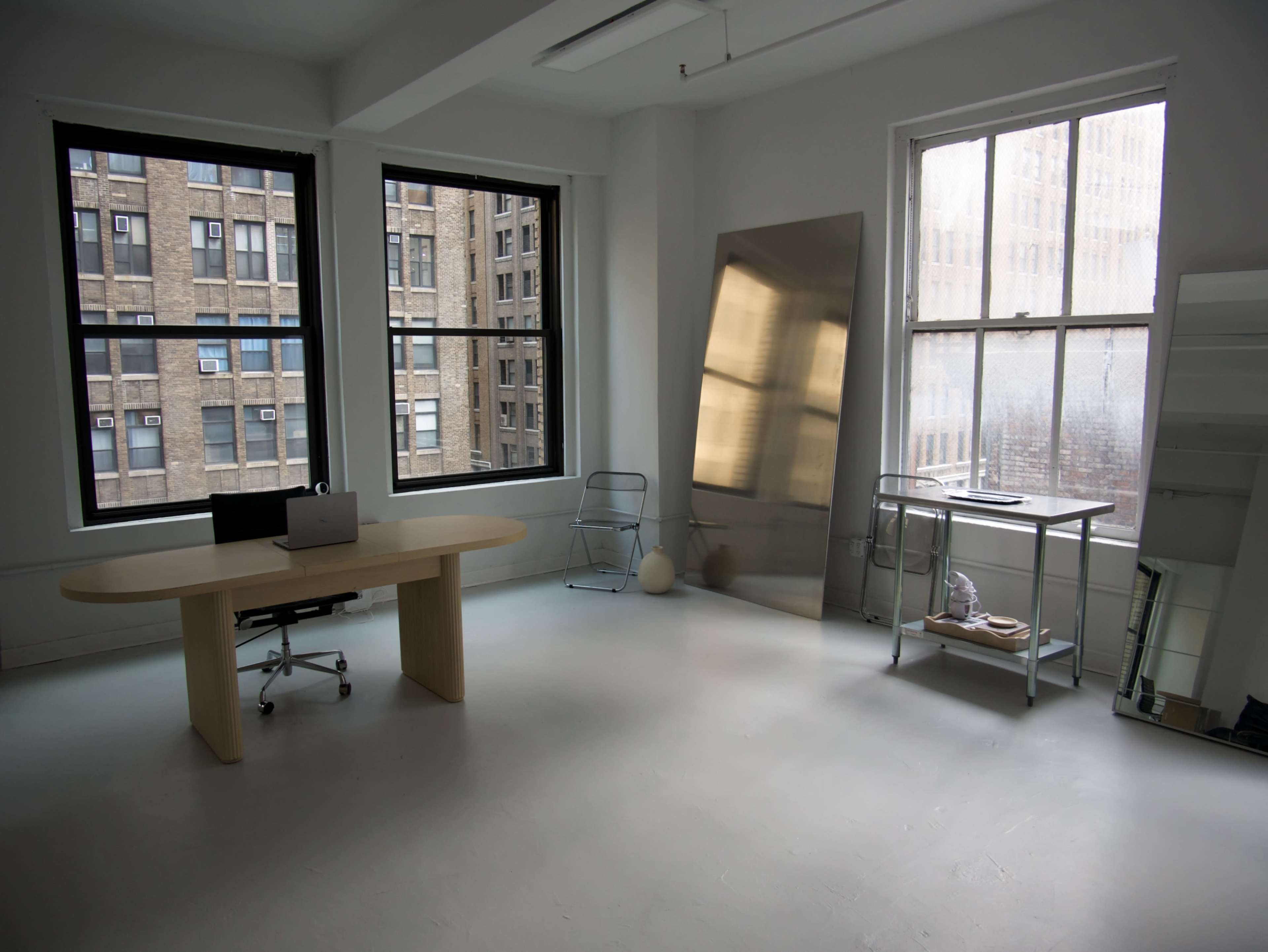 The image shows a sparsely furnished office space with a large desk, a chair, and windows overlooking a brick building exterior.