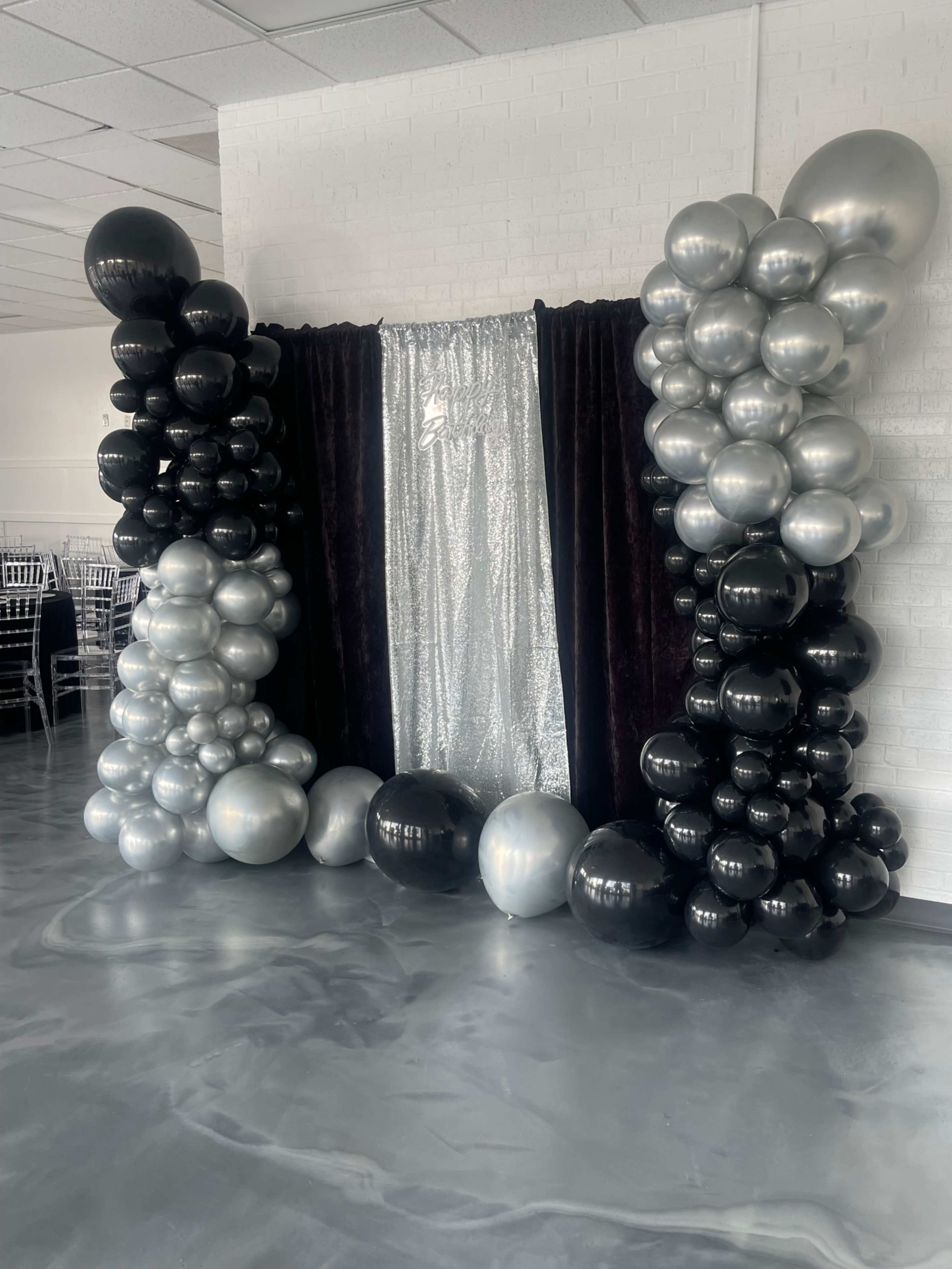 A decorative backdrop for an event, featuring a curtain in the center surrounded by clusters of black and silver balloons.