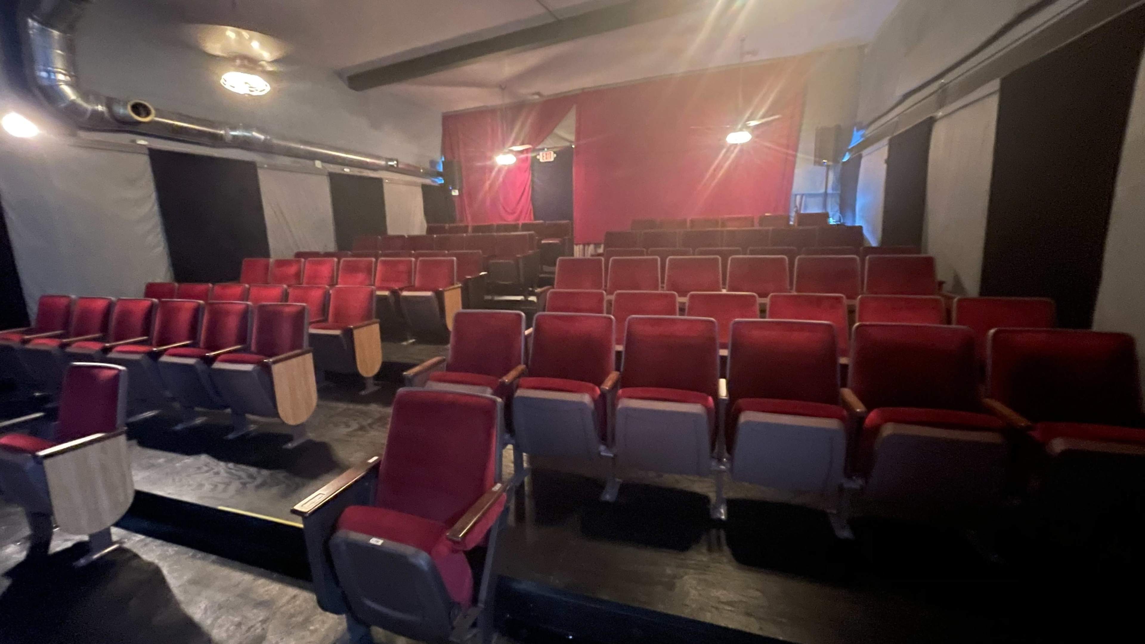 A row of red upholstered theater seats is arranged in a semi-circle facing a stage with dim lighting.