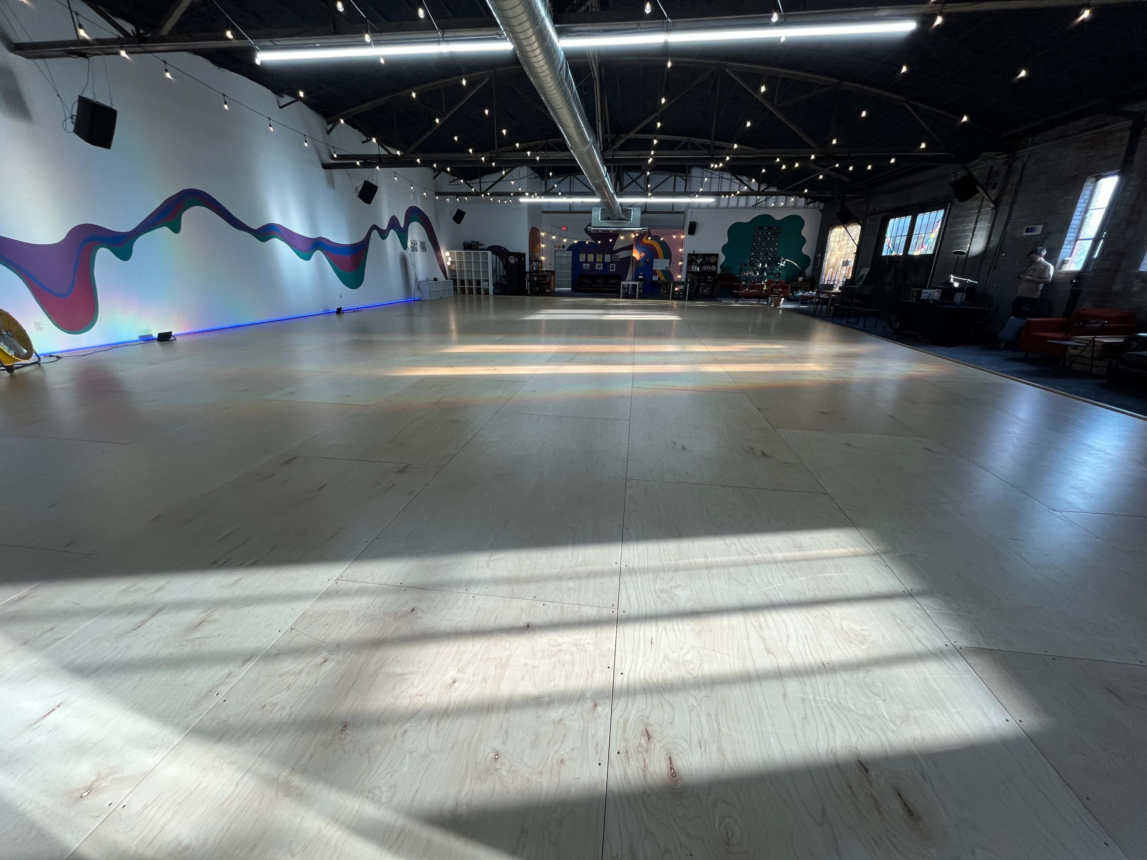 The image shows a spacious indoor area with wooden flooring, illuminated by natural light streaming through large windows, and decorated with colorful wall murals.