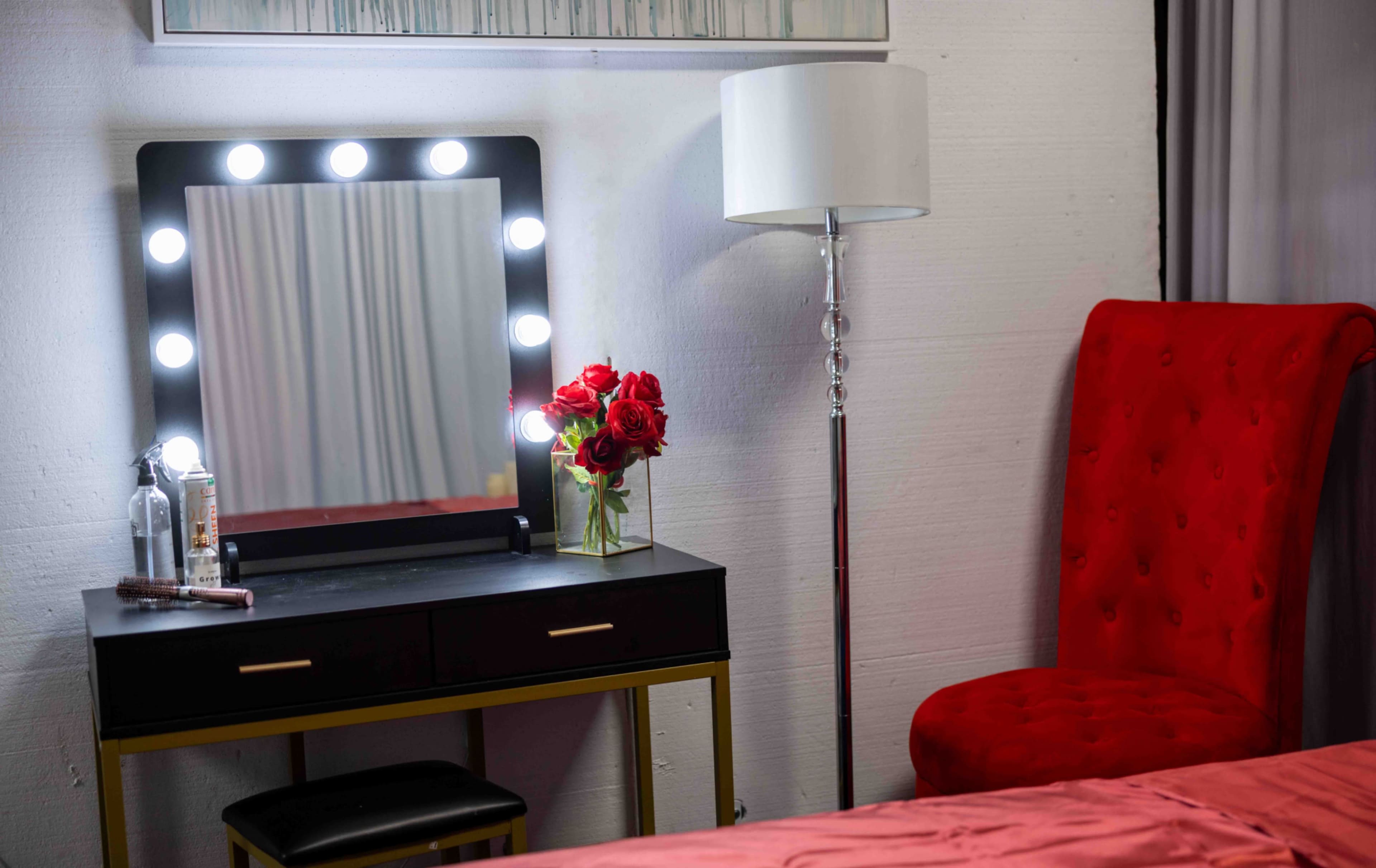 The image features a vanity mirror with lights, a vase of red roses, a floor lamp, and a red upholstered chair next to a bed with a pinkish blanket.