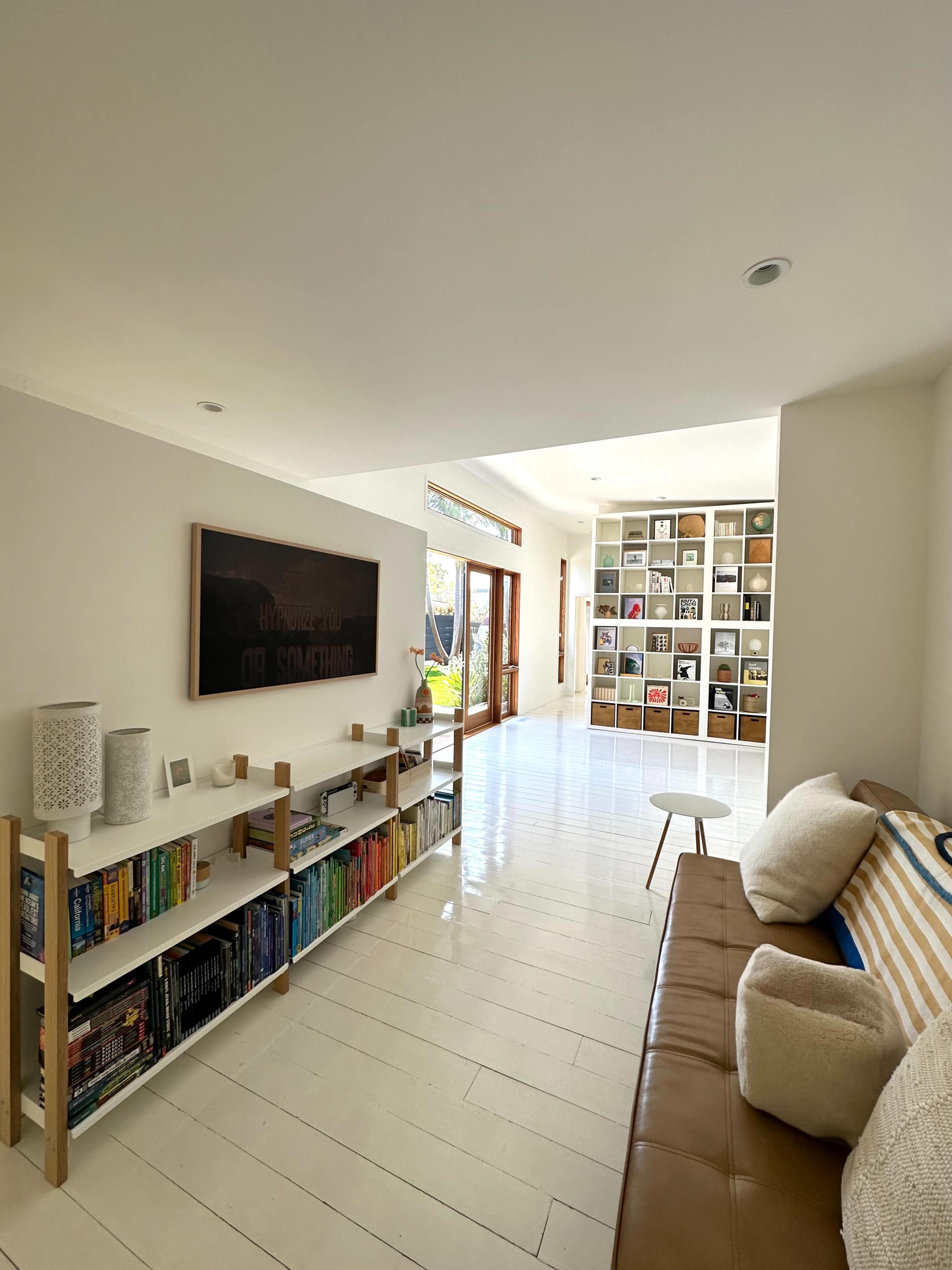The image shows a bright, modern living room featuring a brown leather sofa, a small white table, and a bookshelf filled with colorful books along one wall.