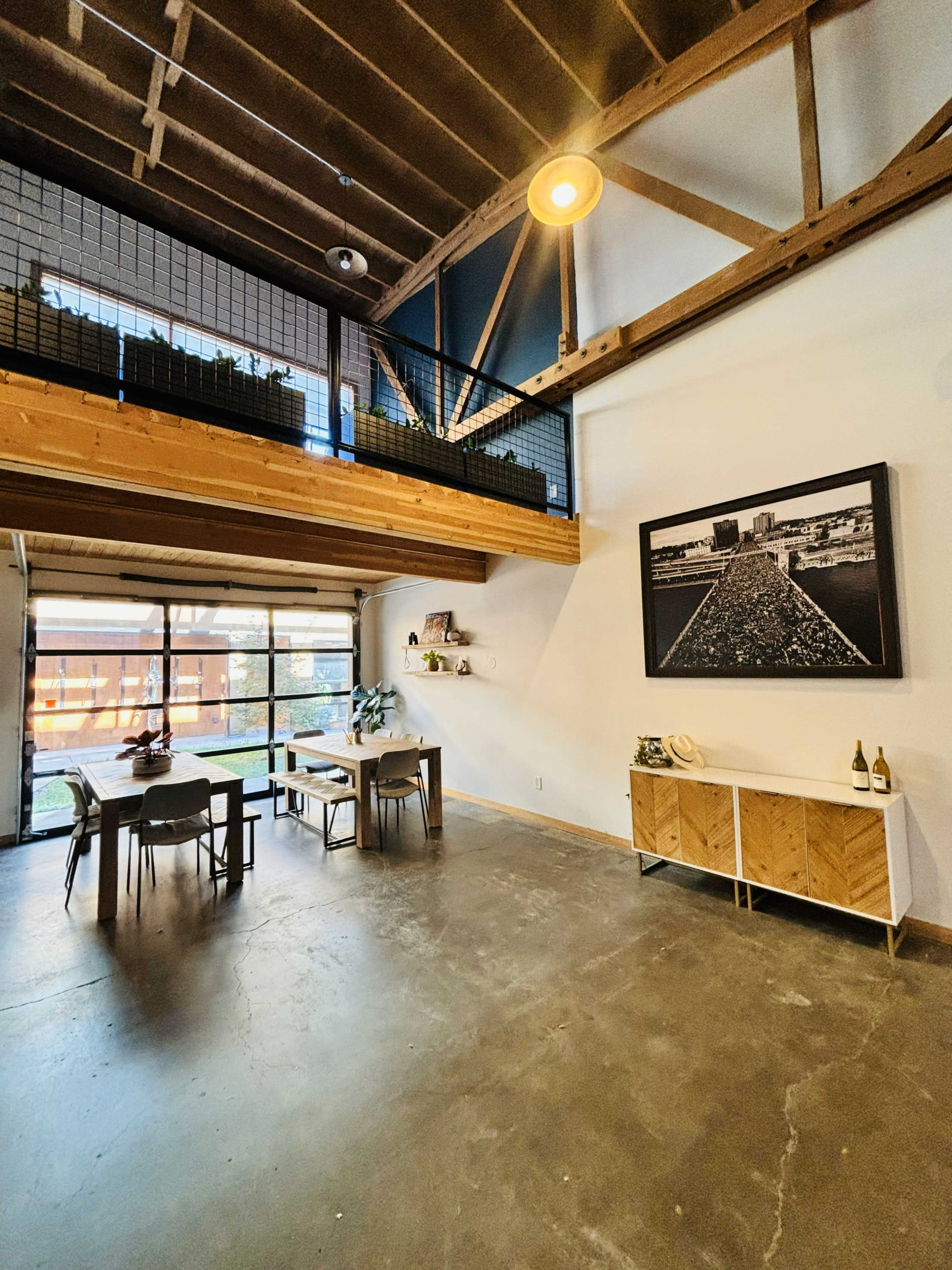 The image shows a spacious interior with a large open area featuring a dining table, a sideboard, and a staircase leading to a lofted space above.