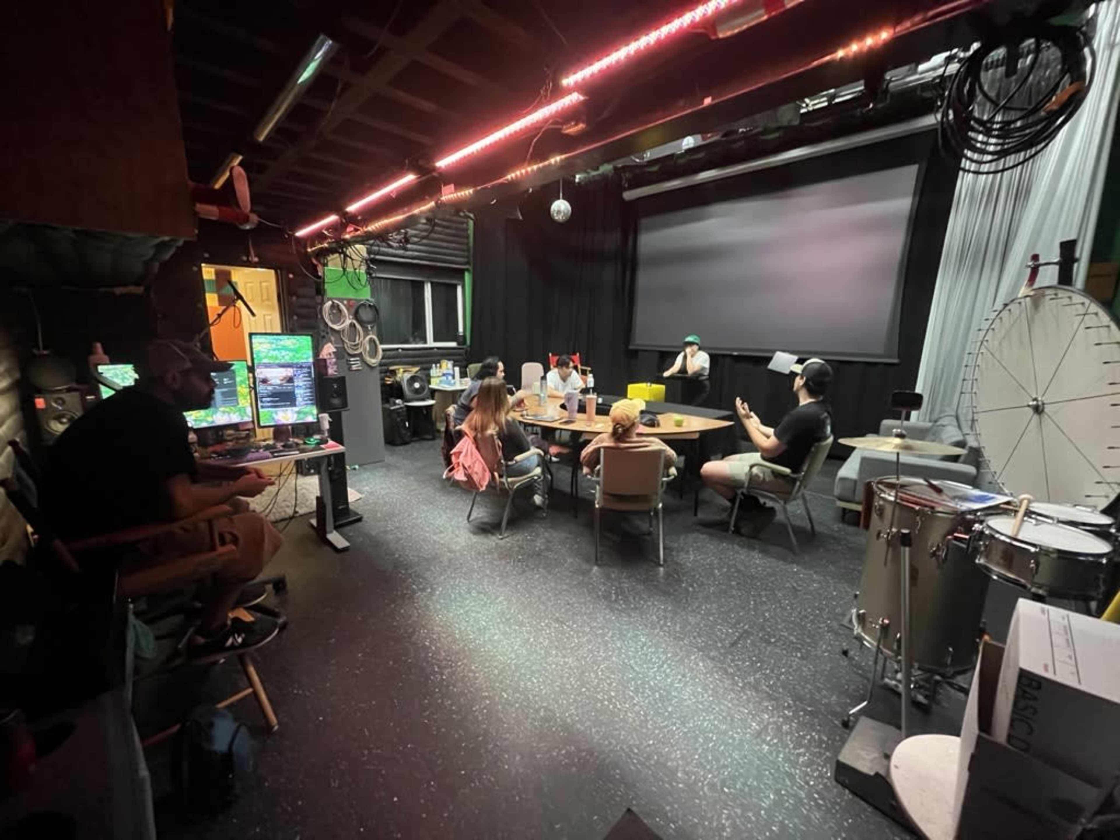 The image shows a dimly lit room with a large screen at the front and several people seated around tables with equipment and monitors set up along the walls.