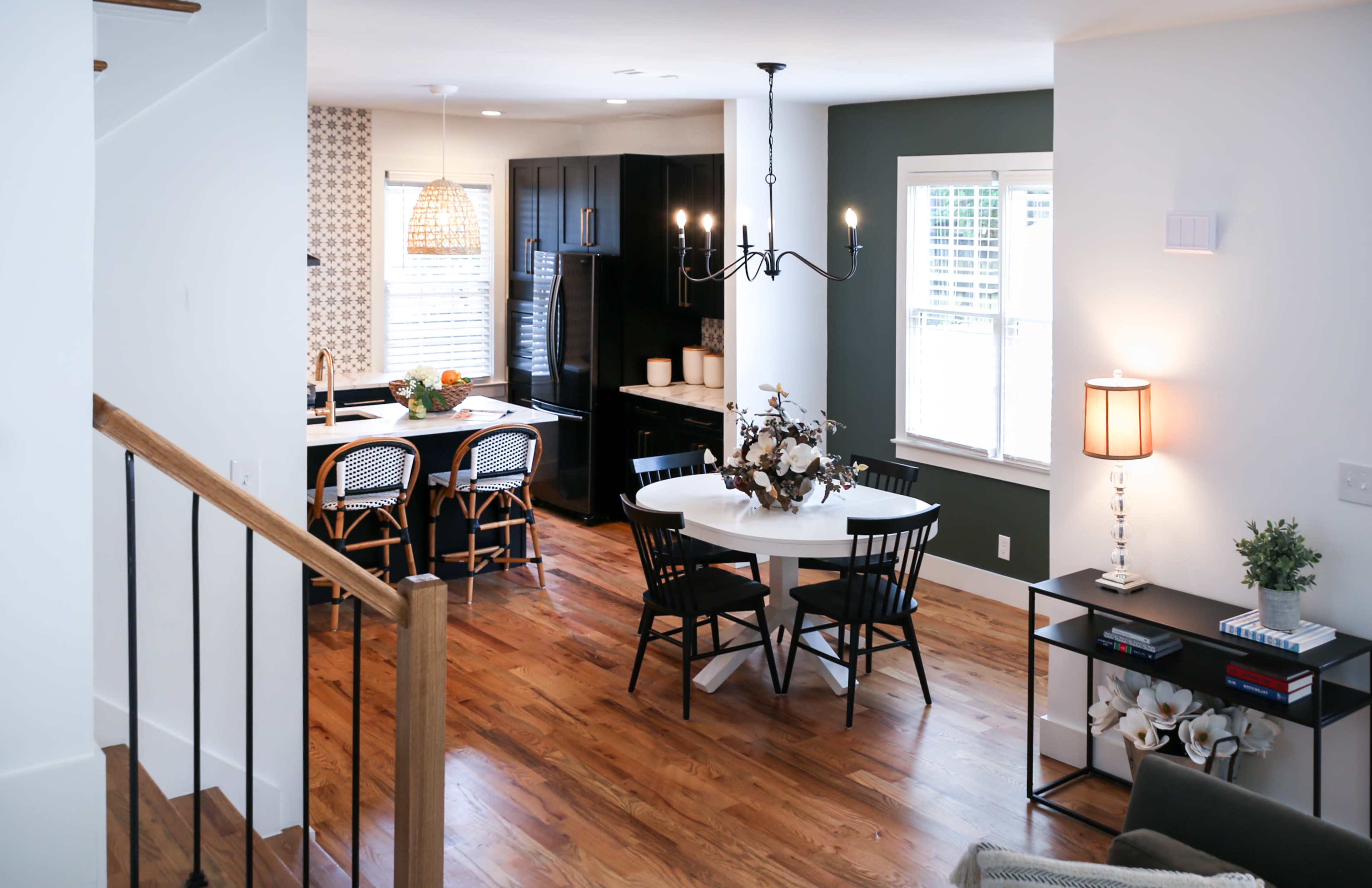 The image shows a modern kitchen and dining area featuring a round white table with black chairs, a dark cabinetry setup, and hardwood flooring.